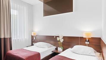 Habitación de hotel con dos camas, cabecera de madera, mesita de noche con lámpara y cortinas traslúcidas.