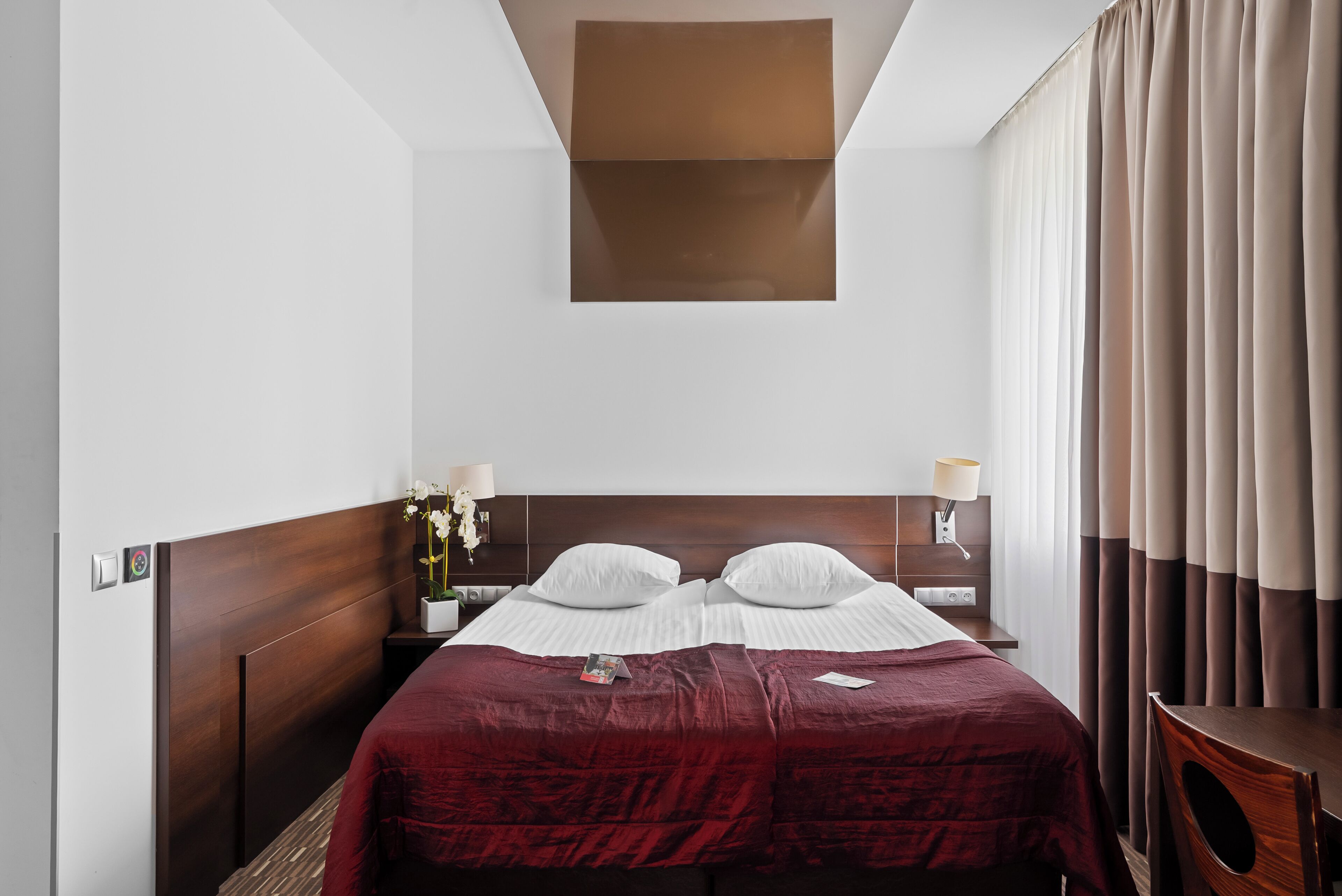 Habitación de hotel con cabecera de madera, una cama grande con colcha roja, dos mesitas de noche con lámparas y vista al cielo a través de una ventana con cortinas.