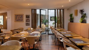 Een moderne eethoek met houten tafels en stoelen, een hoog plafond met dakraam en uitzicht op de omgeving door grote glazen deuren.