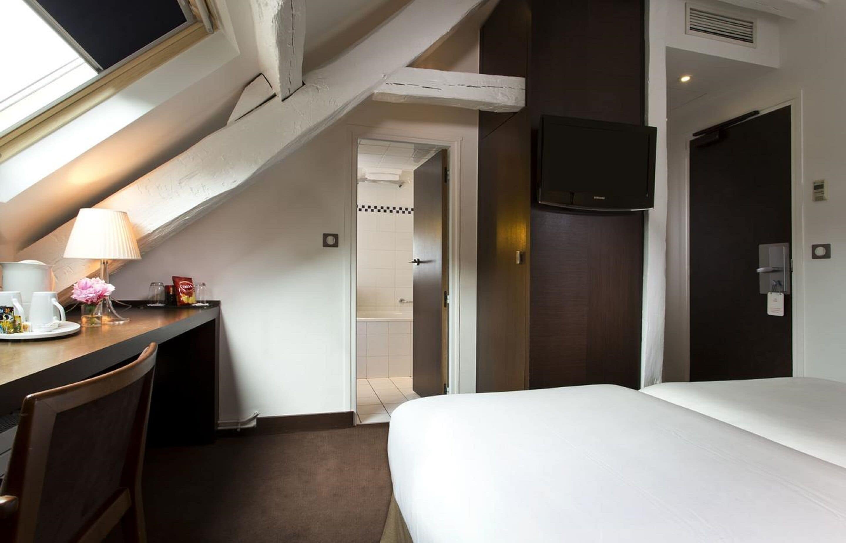 A hotel room with a bed, a desk with a lamp, a TV, and a shower area.