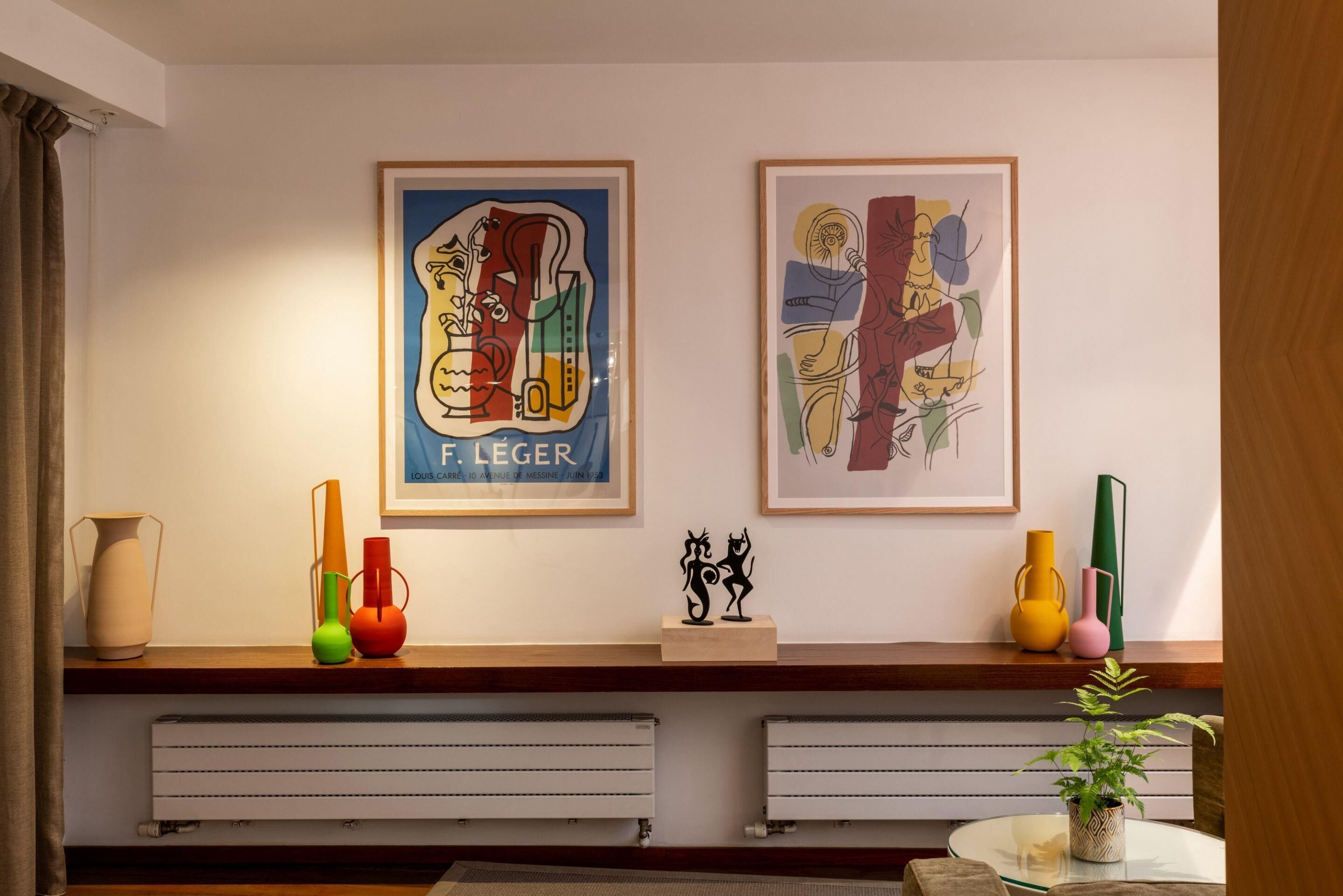 Una habitación con dos cuadros enmarcados en la pared, un estante de madera con jarrones de colores y un radiador debajo.