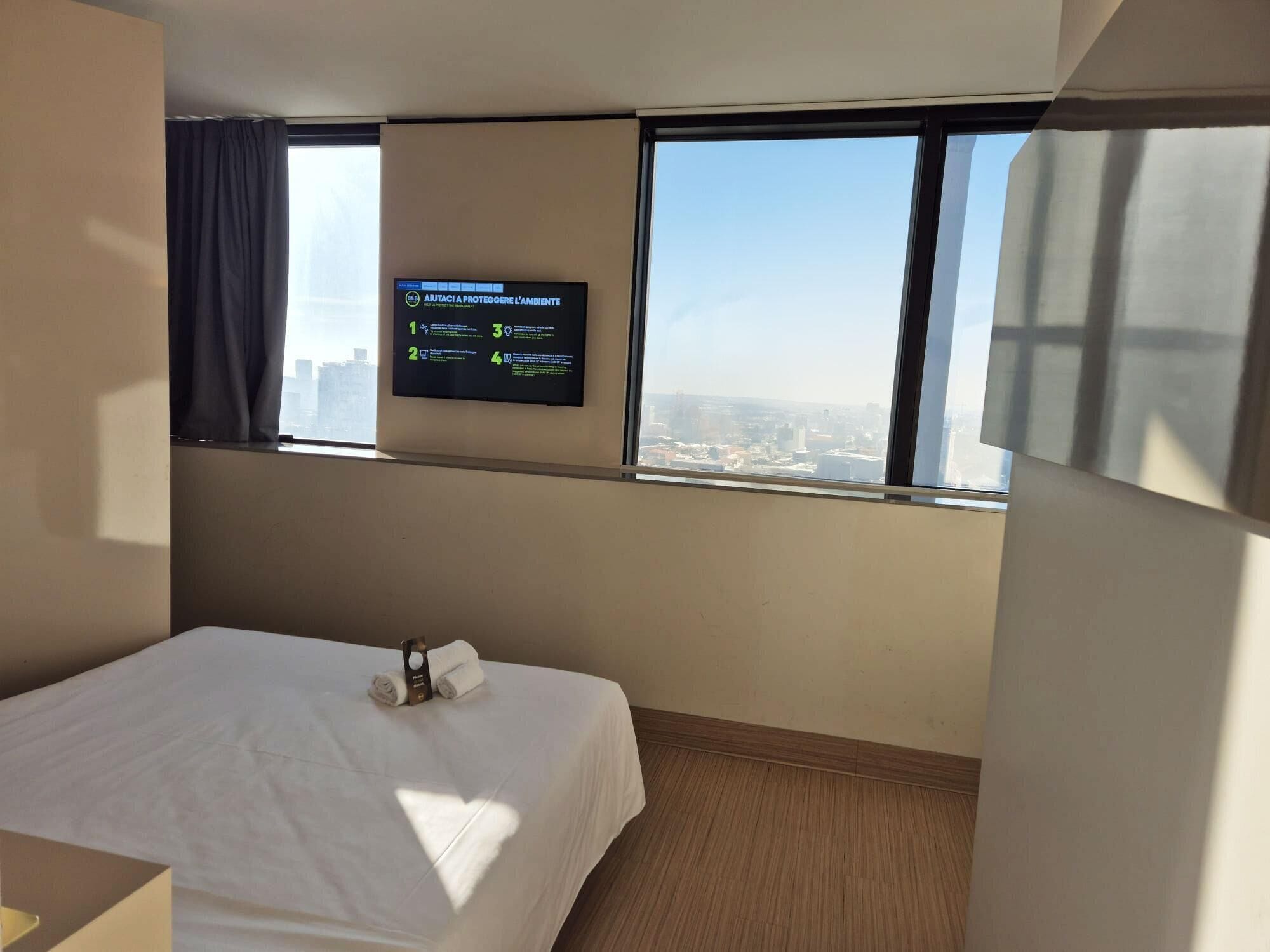 A hotel room with a bed, a TV mounted on the wall displaying a message, and a window with a city view.