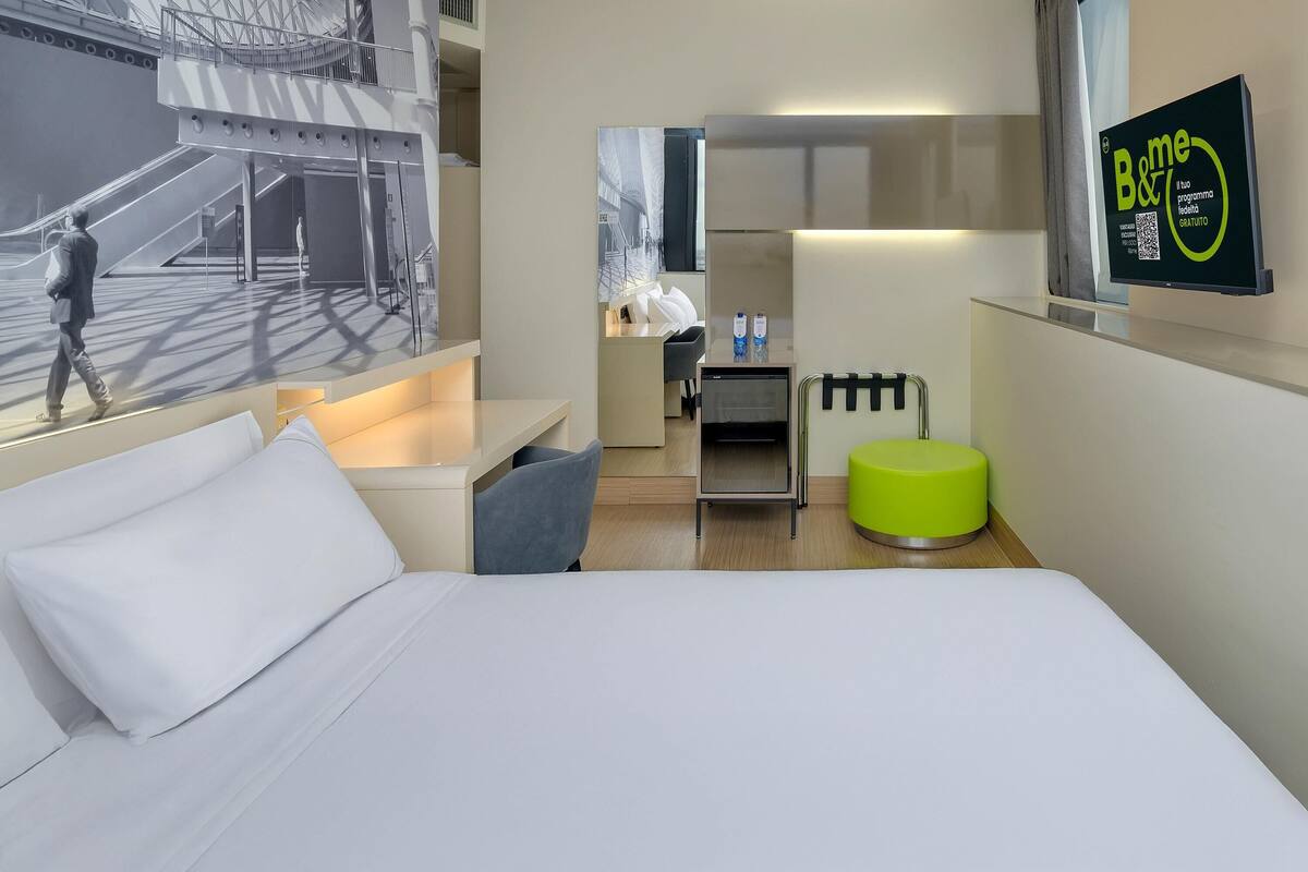 A modern hotel room with a bed, a small seating area, a desk, and a large window.