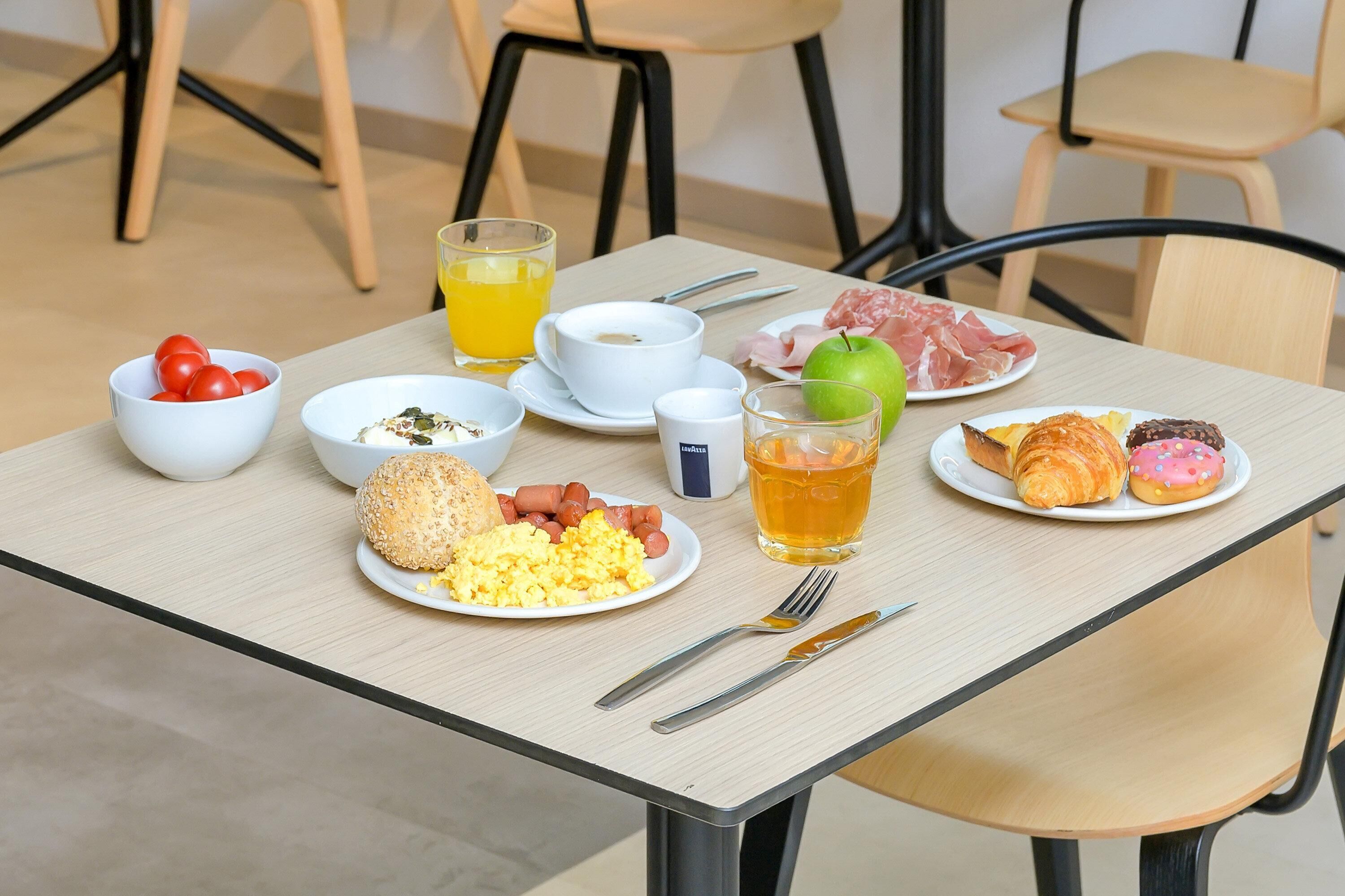 Una mesa de desayuno con platos de huevos, salchichas, tomates, pan, cruasanes y un vaso de jugo de naranja.