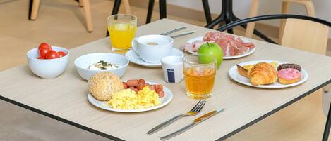 Una mesa de desayuno con platos de huevos, salchichas, tomates, pan, cruasanes y un vaso de jugo de naranja.