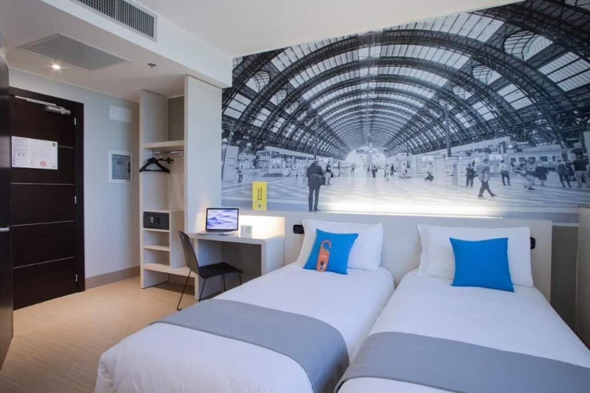 A hotel room with two beds, a desk, a chair, and a large mural of a train station.