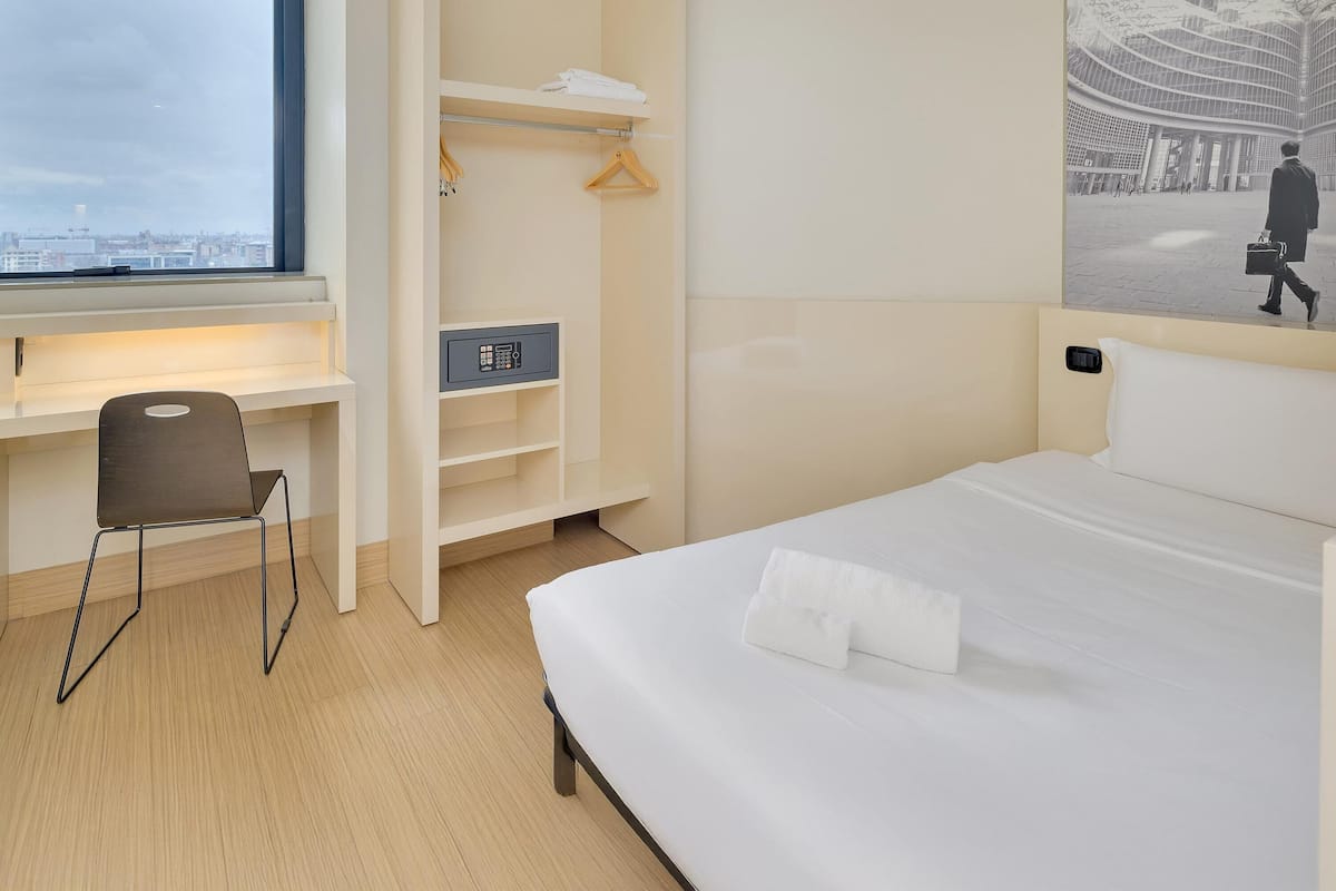A hotel room with a bed, a desk, a chair, a wardrobe, and a large window.