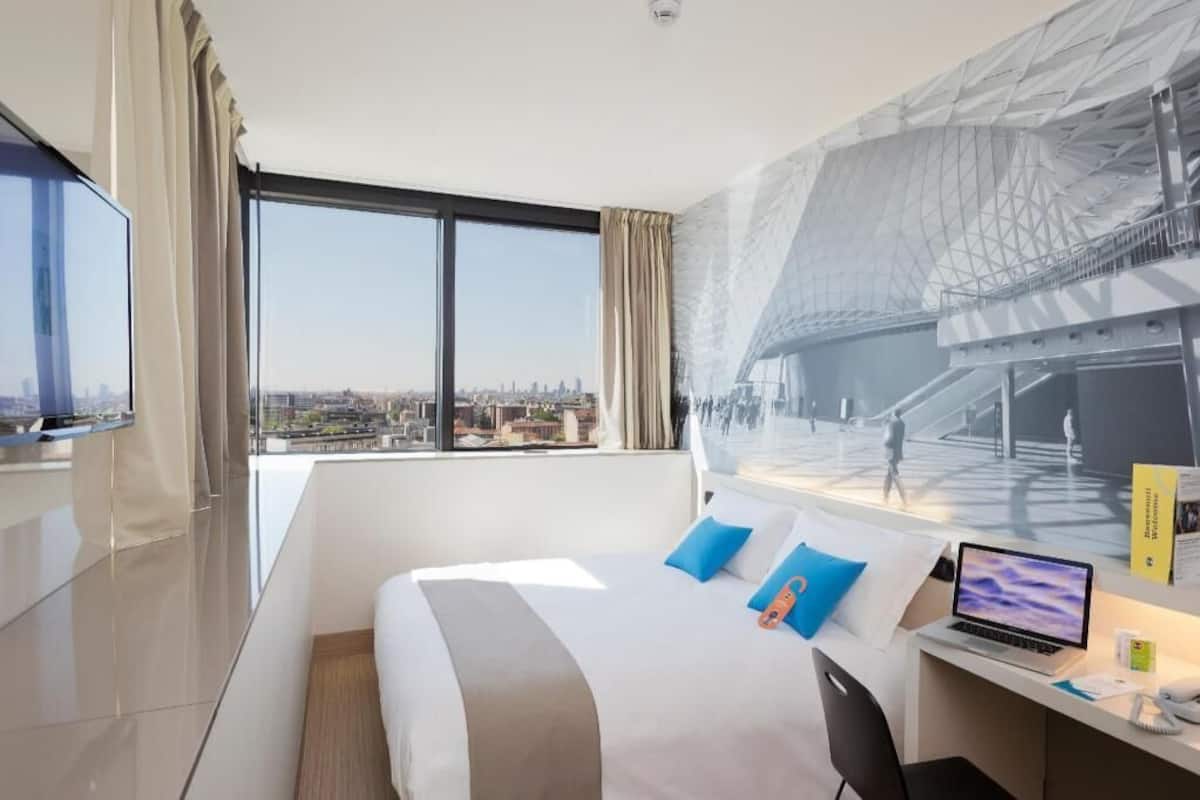 A hotel room with a large bed, a desk with a laptop, a view of a cityscape, and a large window with curtains.