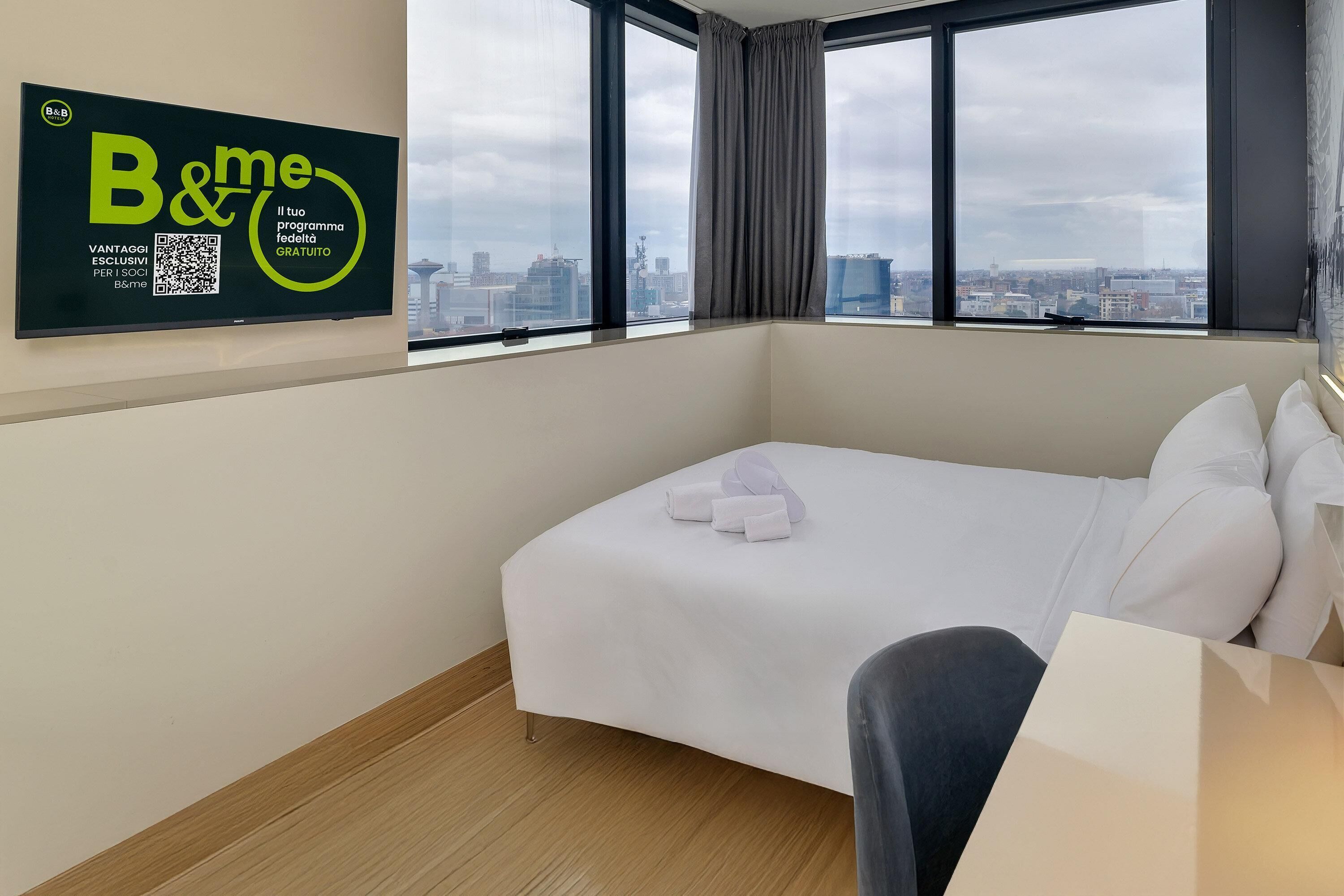 Habitació d'hotel amb un llit, una cadira i una finestra gran que dona a una vista de la ciutat.