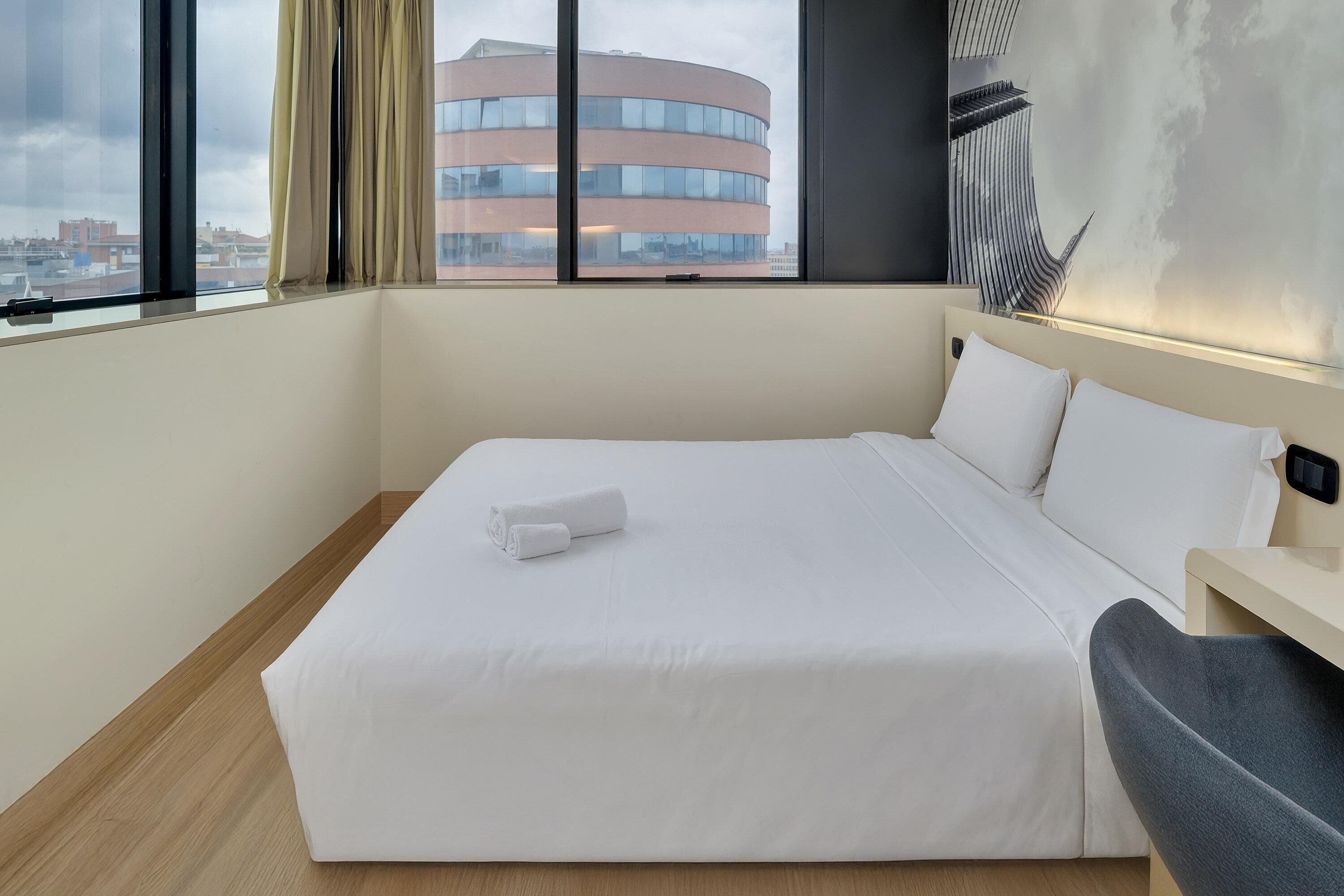 A hotel room with a large bed, a bedside table, and a view of a building through the window.