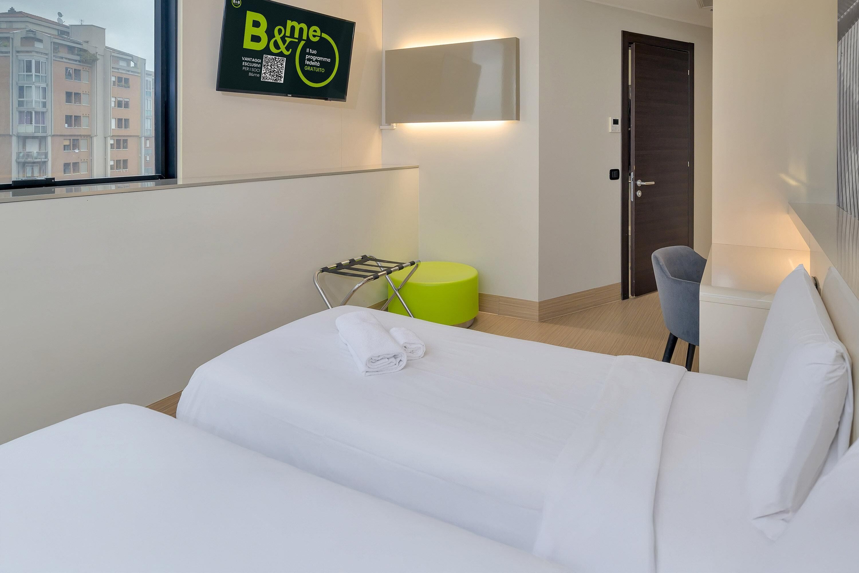 A hotel room with a bed, a green chair, a towel on the bed, a mirror, and a door.