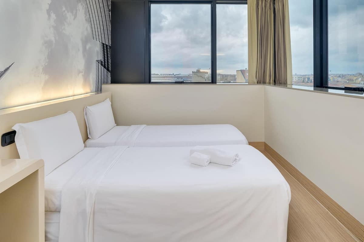 A hotel room with two beds, a large window with a view of the city, and a modern wall design.