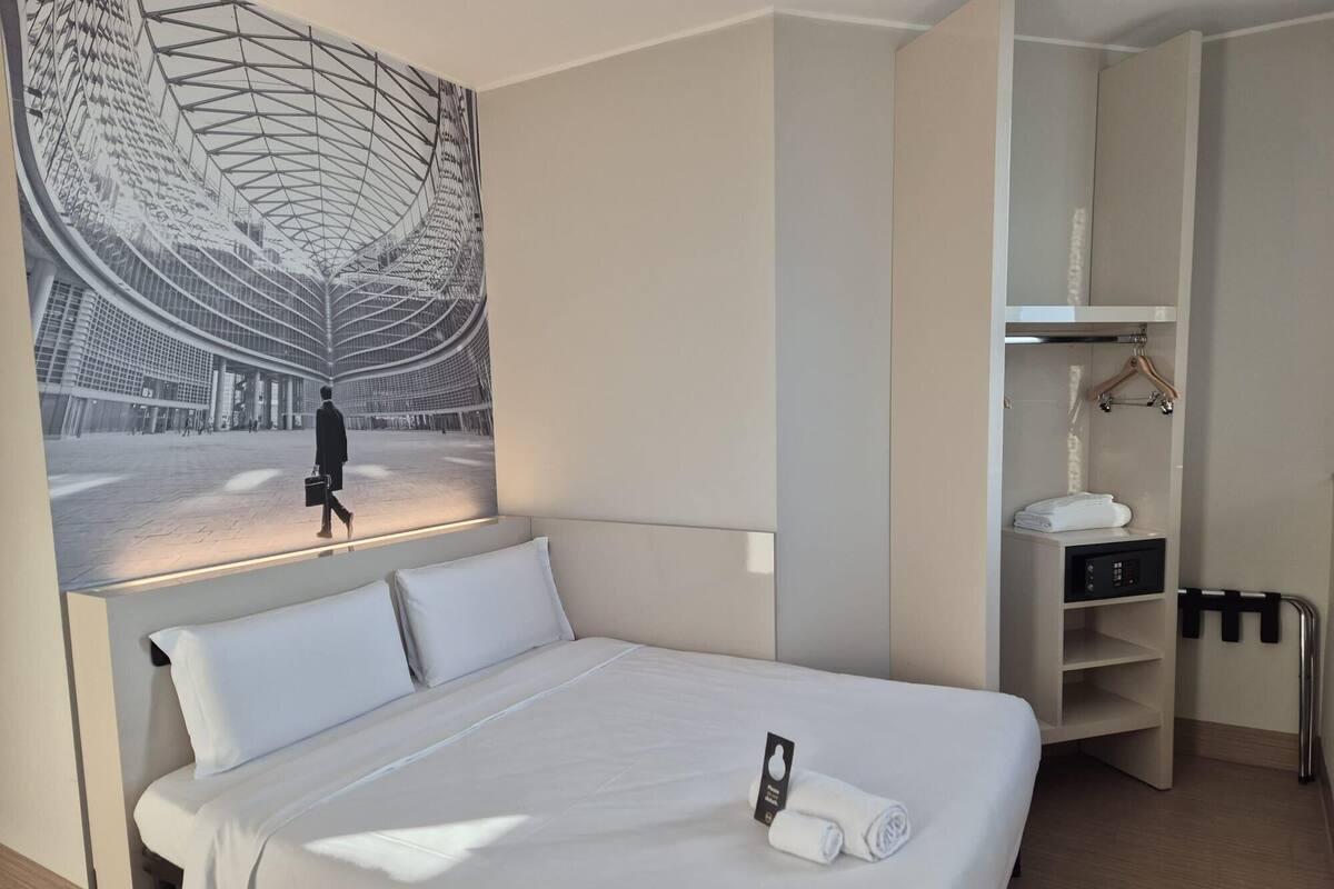 A hotel room with a large bed, a modern abstract painting on the wall, a walk-in closet, and a bathroom with a towel rack and a control panel.