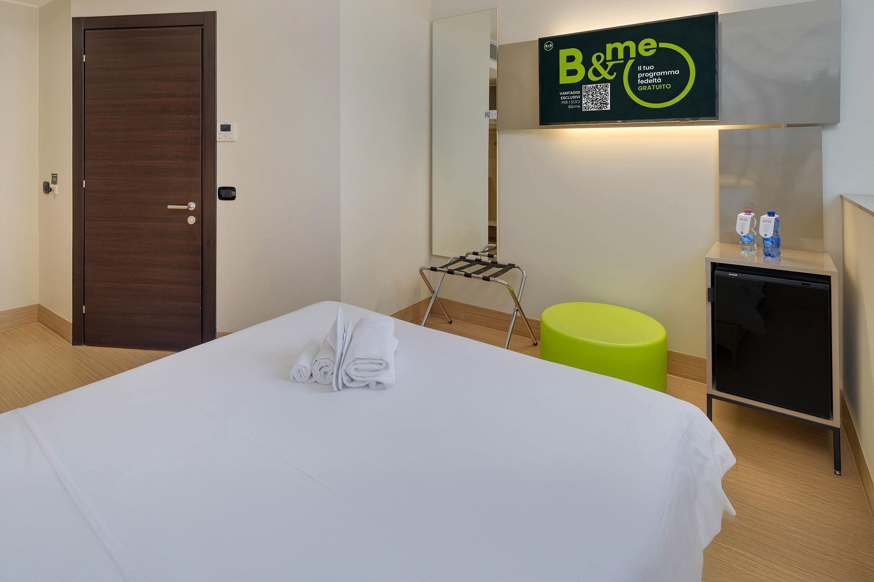 Una habitación de hotel con una cama bien hecha, una mesita con una toalla, un taburete verde, un minibar y un cartel de B&me en la pared.