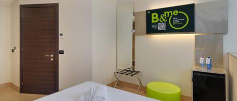 Una habitación de hotel con una cama bien hecha, una mesita con una toalla, un taburete verde, un minibar y un cartel de B&me en la pared.