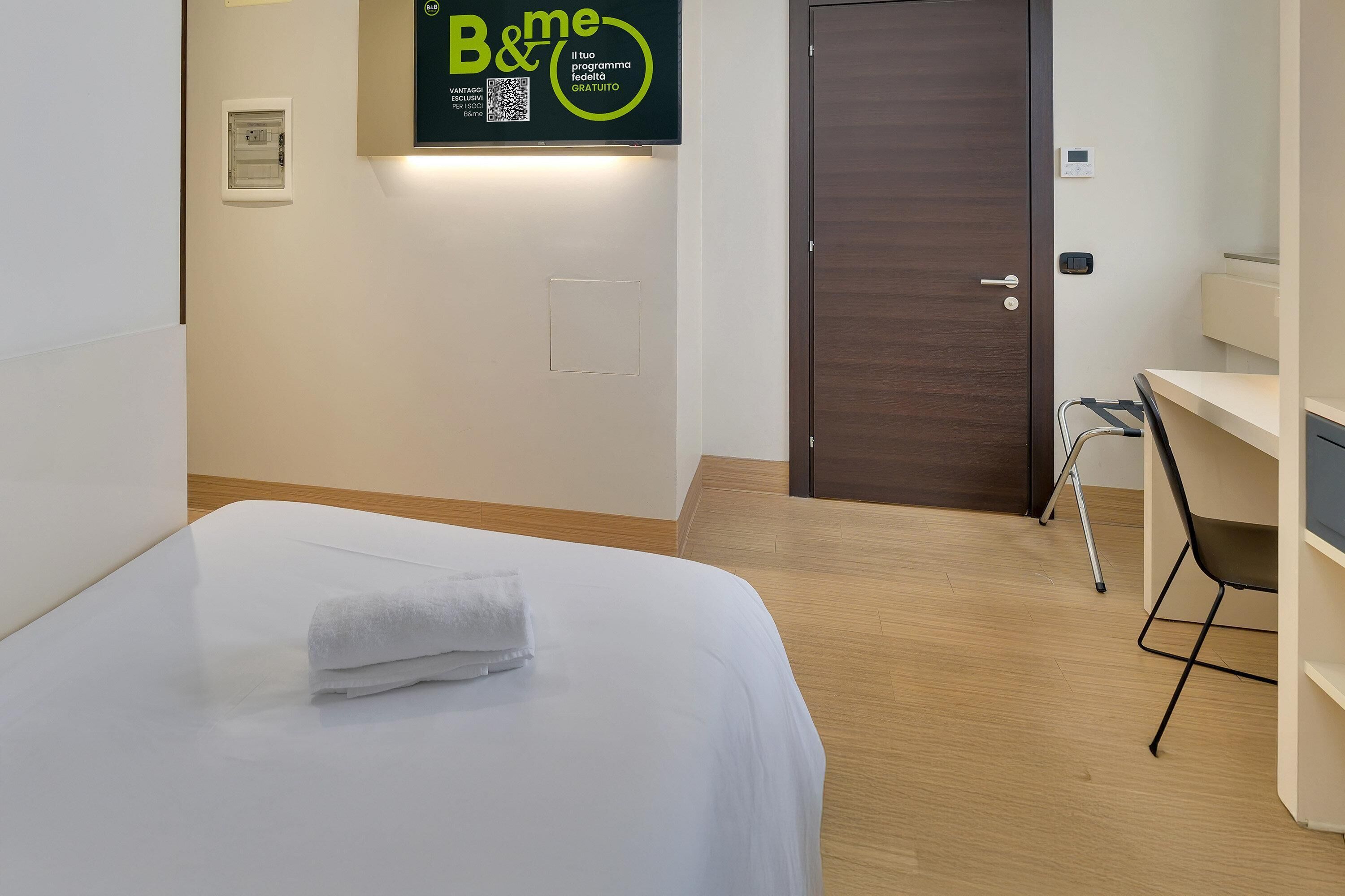 A hotel room with a bed, a desk, a chair, a door, and a wall-mounted TV displaying a program.