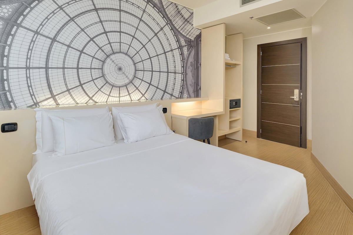 A hotel room with a large bed, a desk, a wardrobe, and a wall mural.