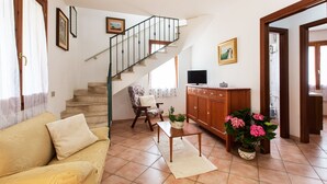 Una sala de estar con un sofá, una mesa de centro, un televisor sobre un mueble de madera, una escalera y una puerta que da a otra habitación.