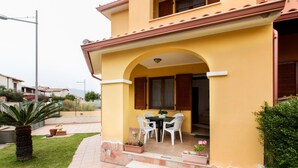 Un edificio amarillo con un patio techado que incluye una mesa, sillas, escalones de piedra y plantas en macetas.