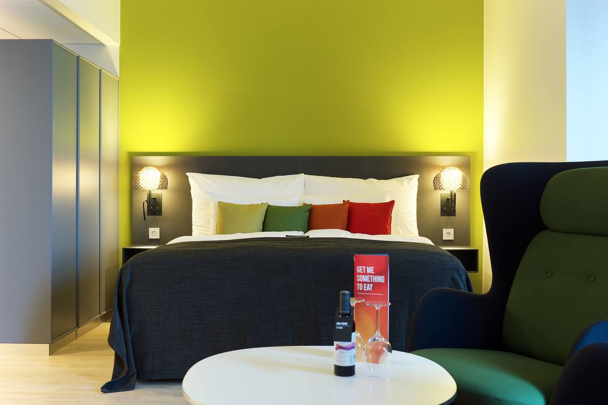 A hotel room with a bed, bedside lamps, a table with a bottle and glass, and a red sign on the wall.