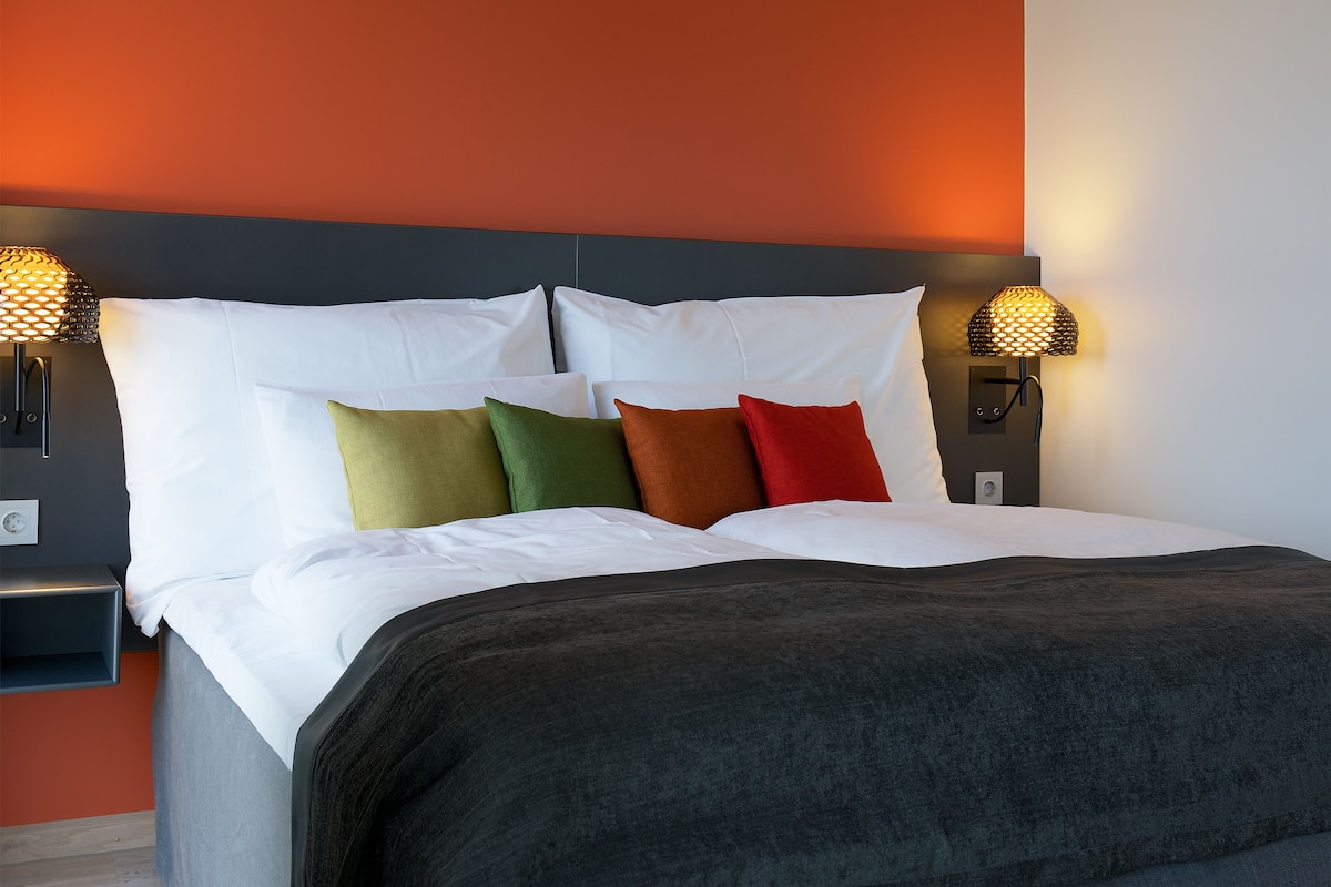 A neatly made bed with white linens and multiple pillows in various colors. There are two wall-mounted lamps on either side of the bed.