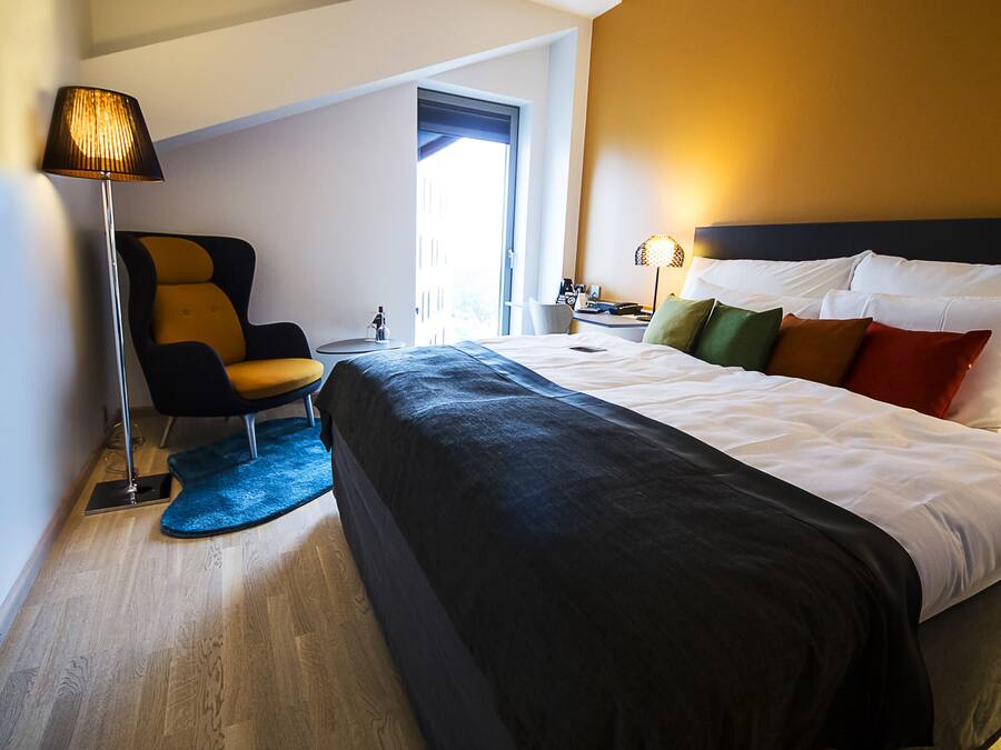 A modern hotel room with a large bed, a chair, a desk, and a lamp.