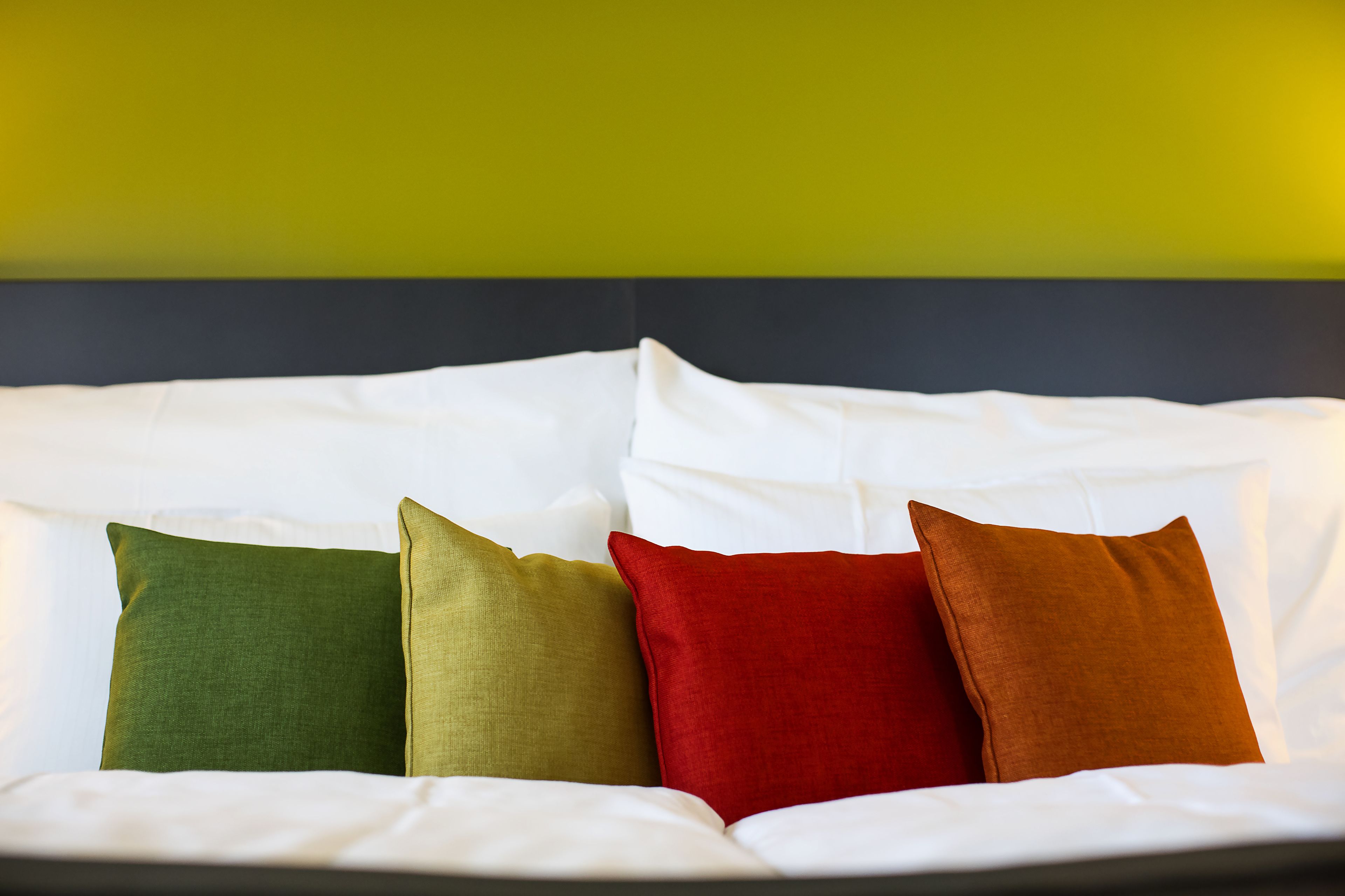 Una cama con ropa blanca y cuatro almohadas en tonos verde, oliva, rojo y marrón.