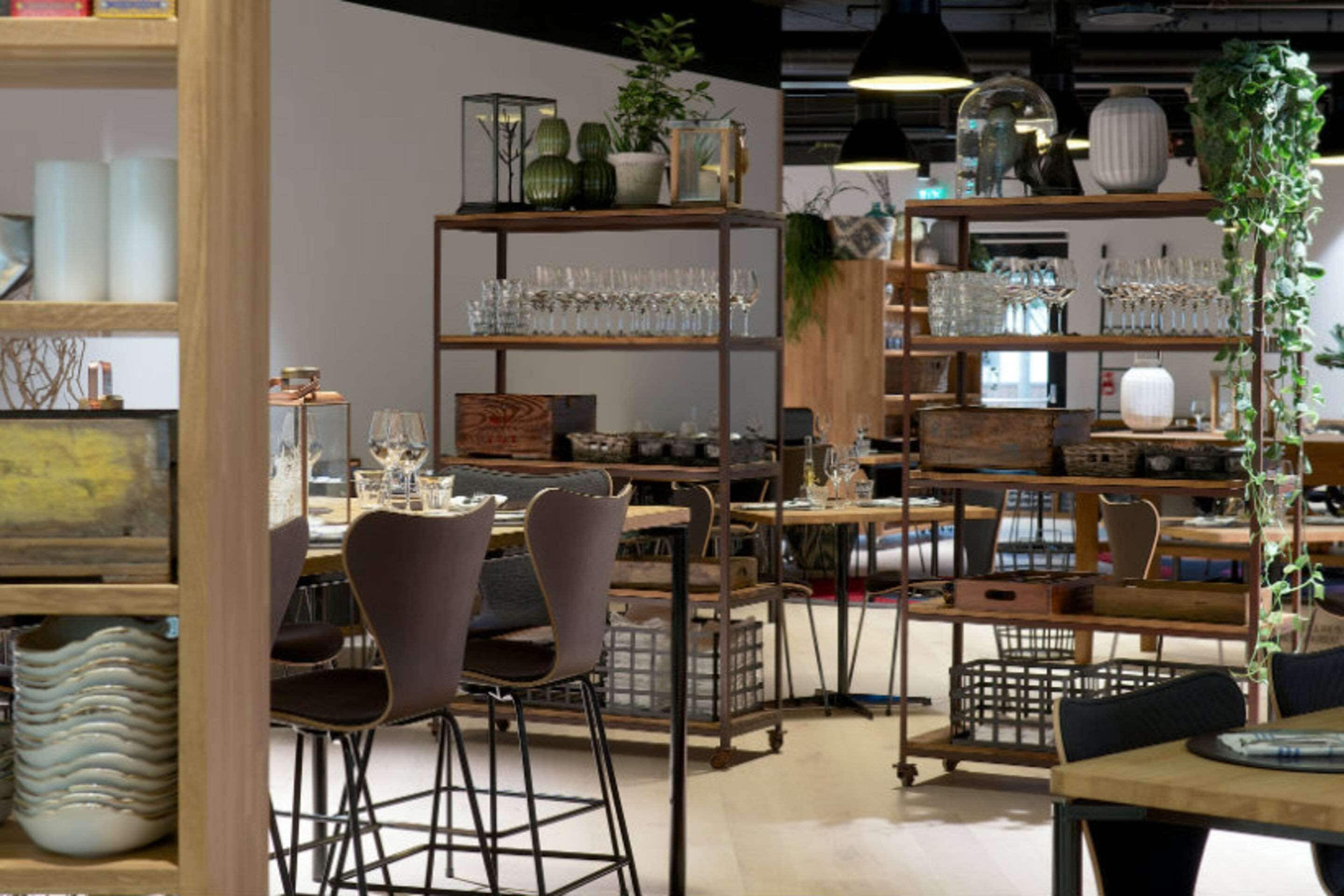 A restaurant interior with wooden shelves displaying glasses and dishes, a dining area with chairs, and a potted plant.