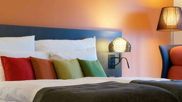 Una habitación de hotel con una cama bien hecha, almohadas coloridas, una lámpara de pared y una silla.
