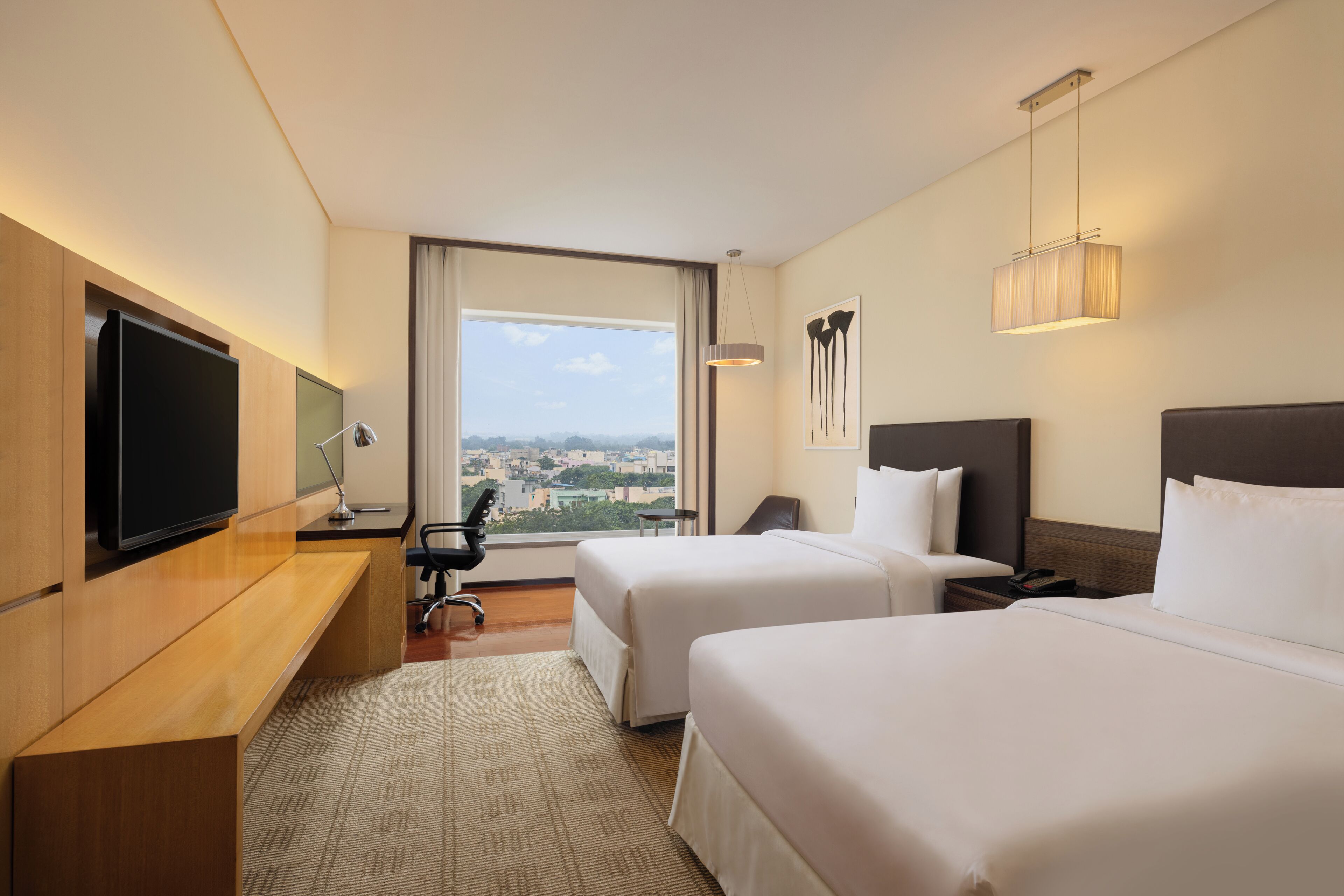 A hotel room with two beds, a desk, a TV, and a large window offering a city view.