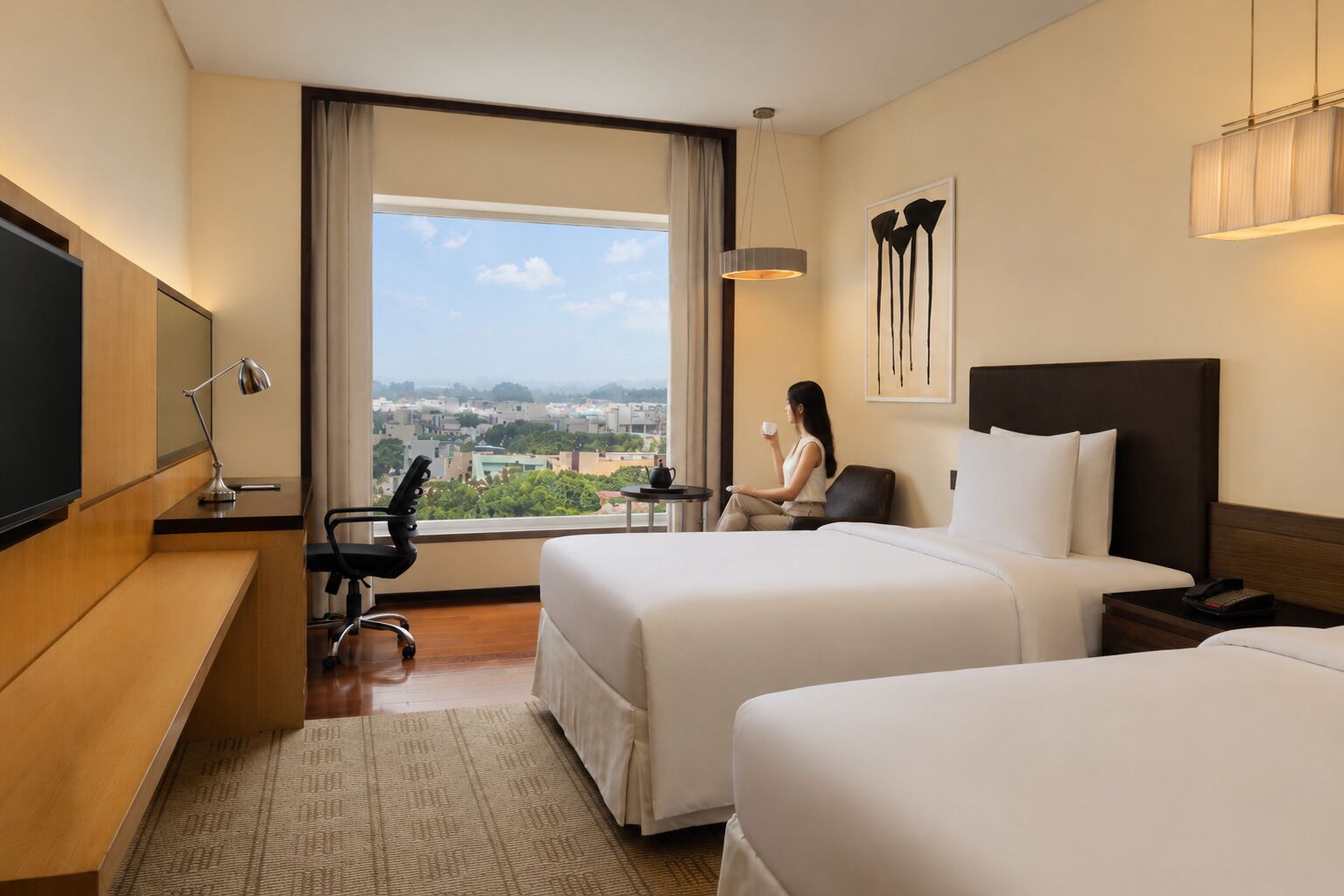 A hotel room with two beds, a desk, a chair, a TV, and a large window with a city view.