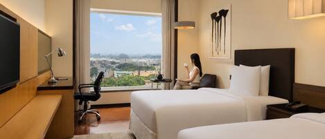 A hotel room with two beds, a desk, a chair, a TV, and a large window with a city view.