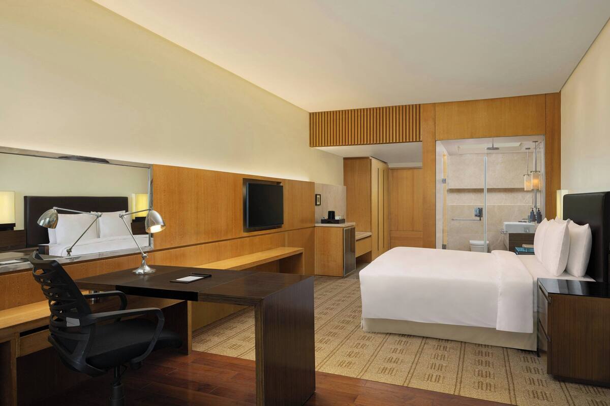 A hotel room with a bed, desk, chair, TV, and a bathroom with a shower and sink.