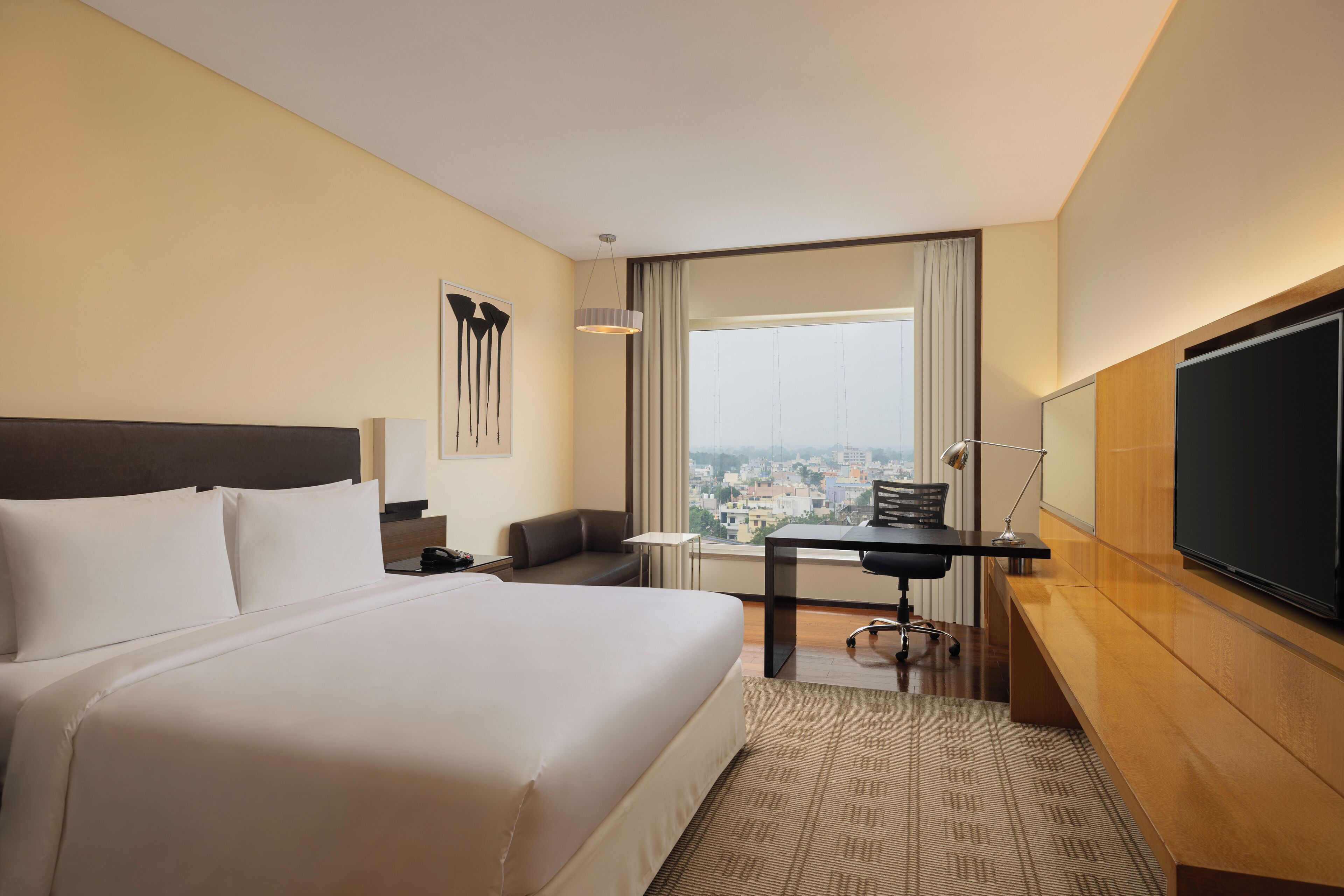 Una habitación de hotel con una cama grande, un escritorio, una silla, un televisor y vistas a un paisaje urbano a través de la ventana.
