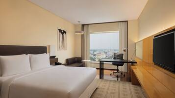 Una habitación de hotel con una cama grande, un escritorio, una silla, un televisor y vistas a un paisaje urbano a través de la ventana.