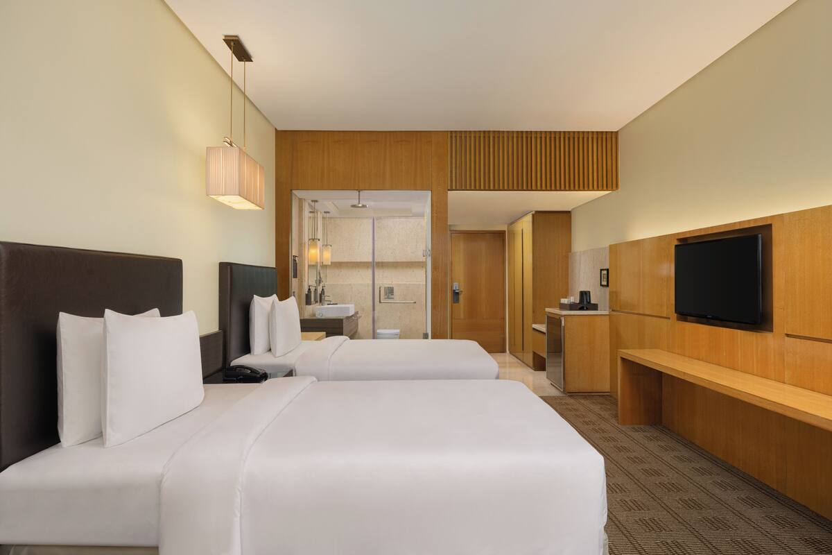 A hotel room with two beds, a TV, a desk, and a bathroom visible through an open door.