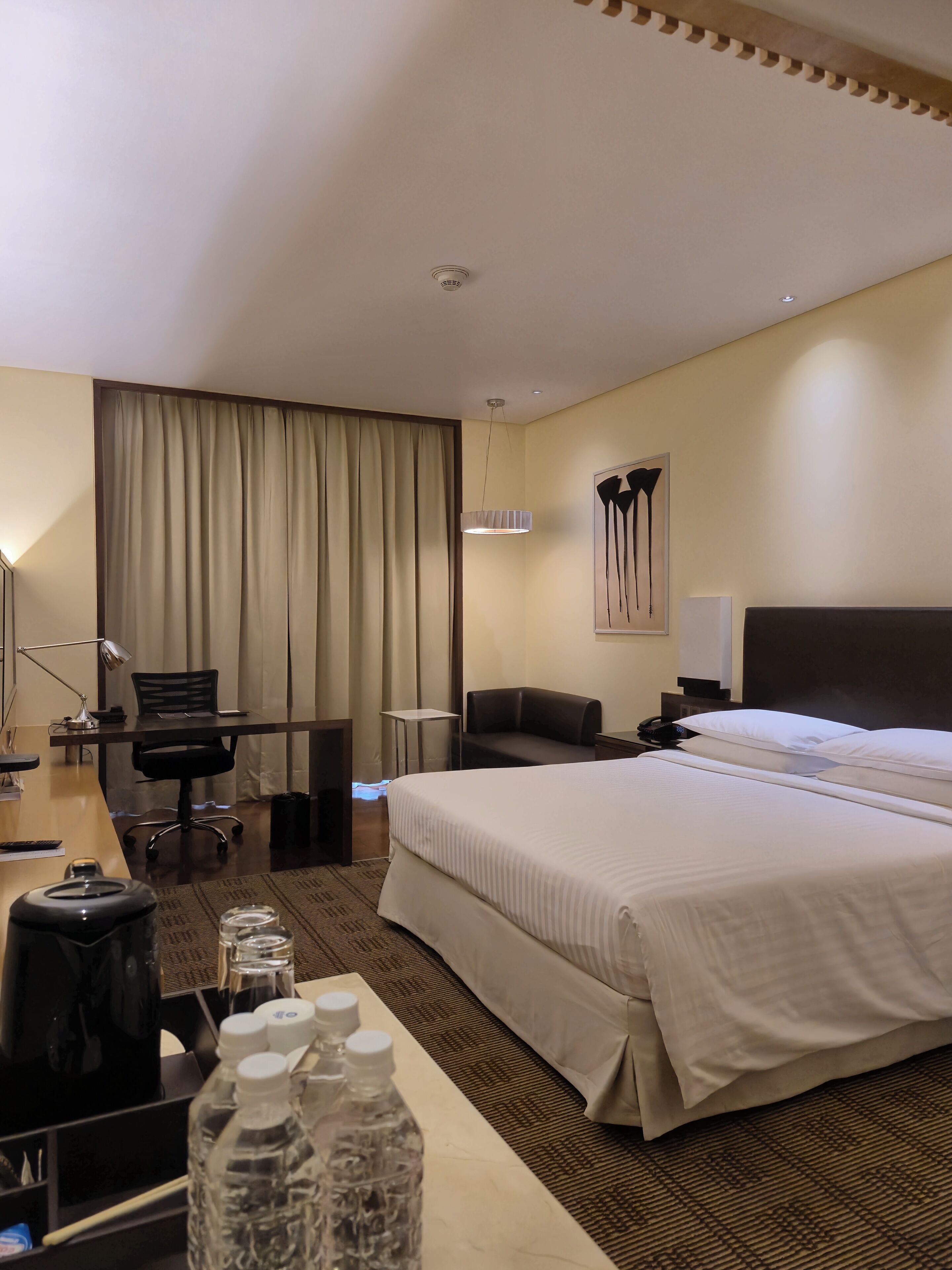 A hotel room with a large bed, a desk with a chair, a small table, and a lamp. There is a curtain on the window and a painting on the wall.