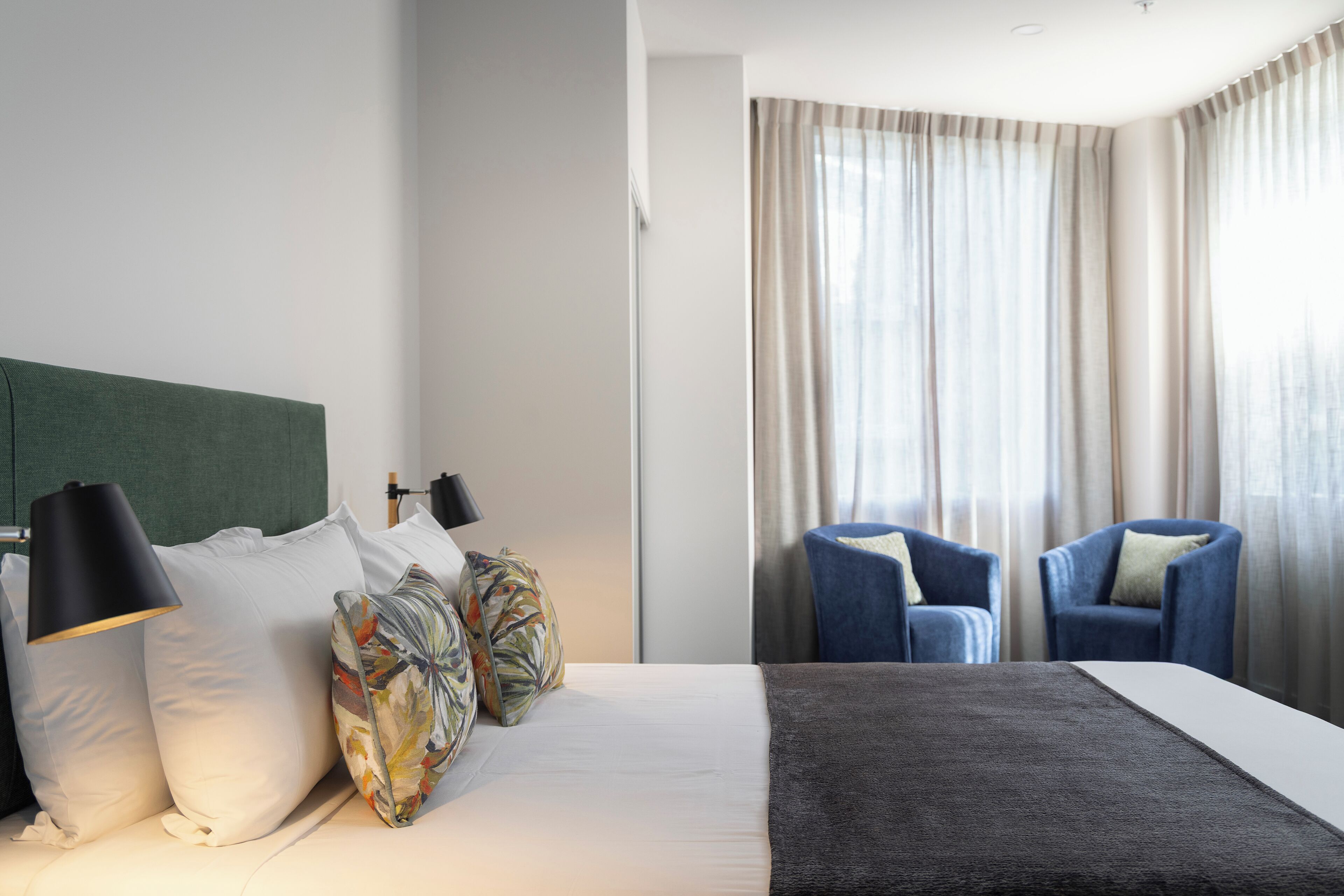 A hotel room with a bed, a headboard, a lamp, two chairs, and a window with curtains.