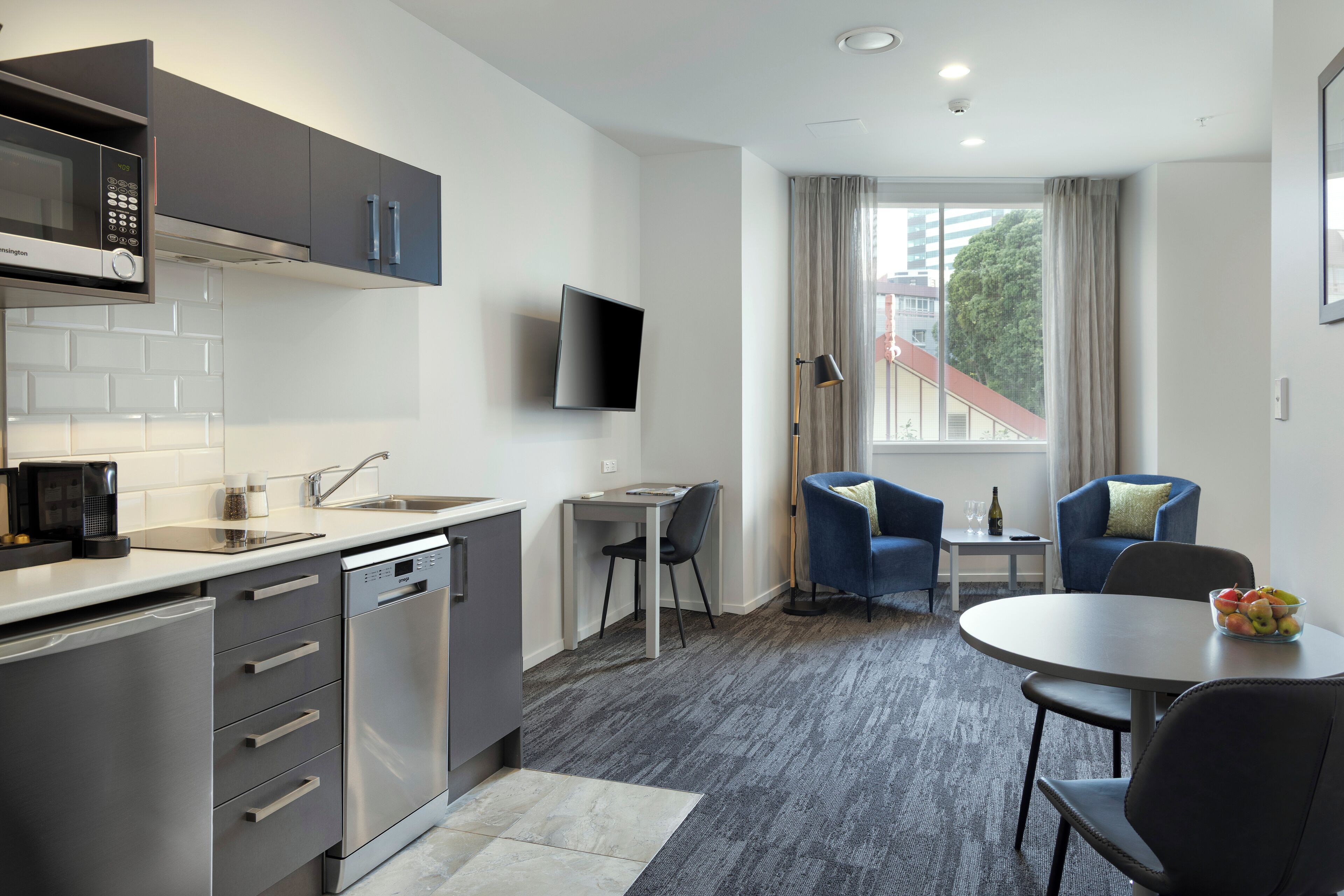 A modern kitchen with a built-in microwave, dishwasher, and a flat-screen TV. There is a dining area with a round table and two chairs, and a window with curtains.