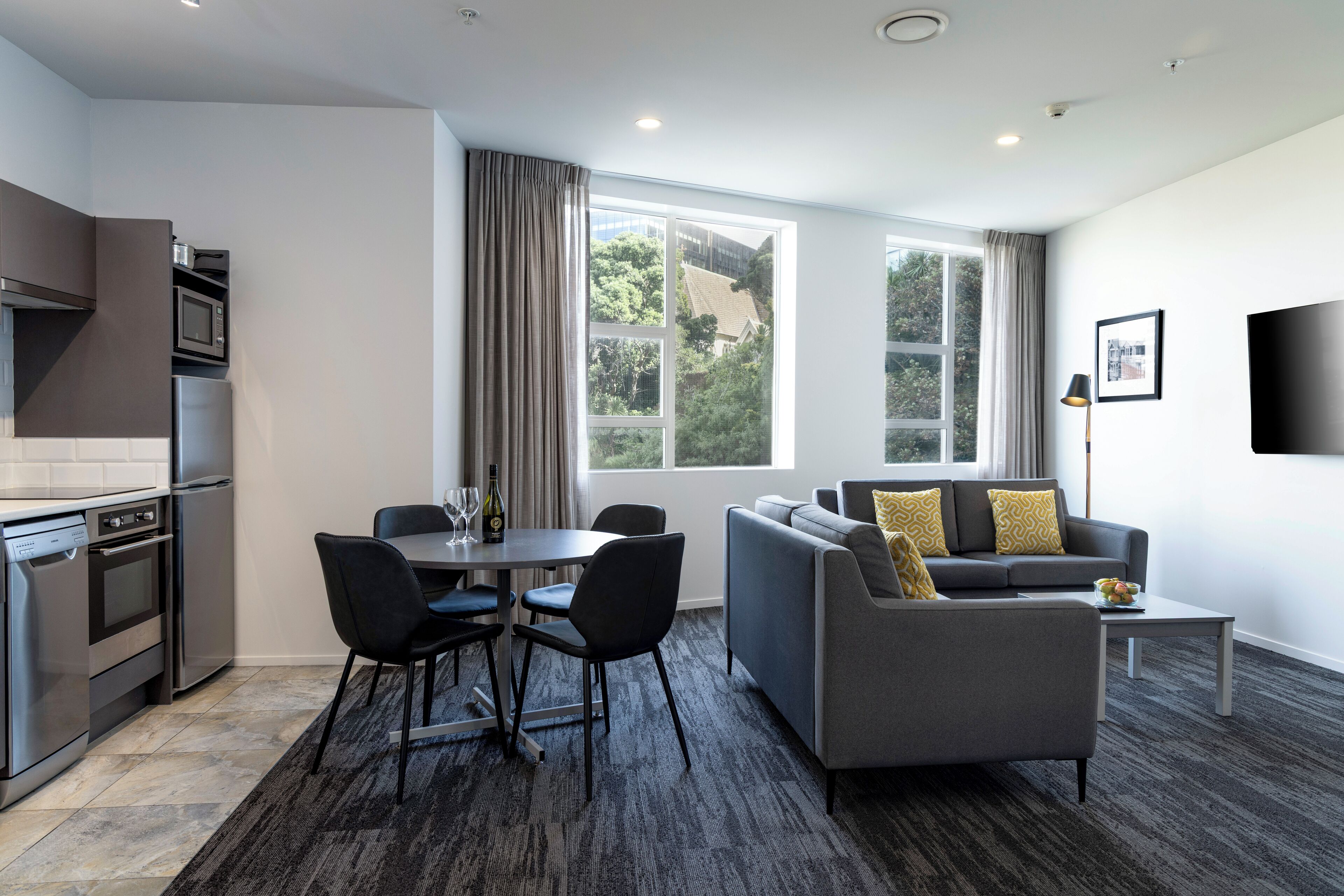 A modern living room with a grey sofa, black chairs, a dining table, and a kitchen area with a microwave and oven.