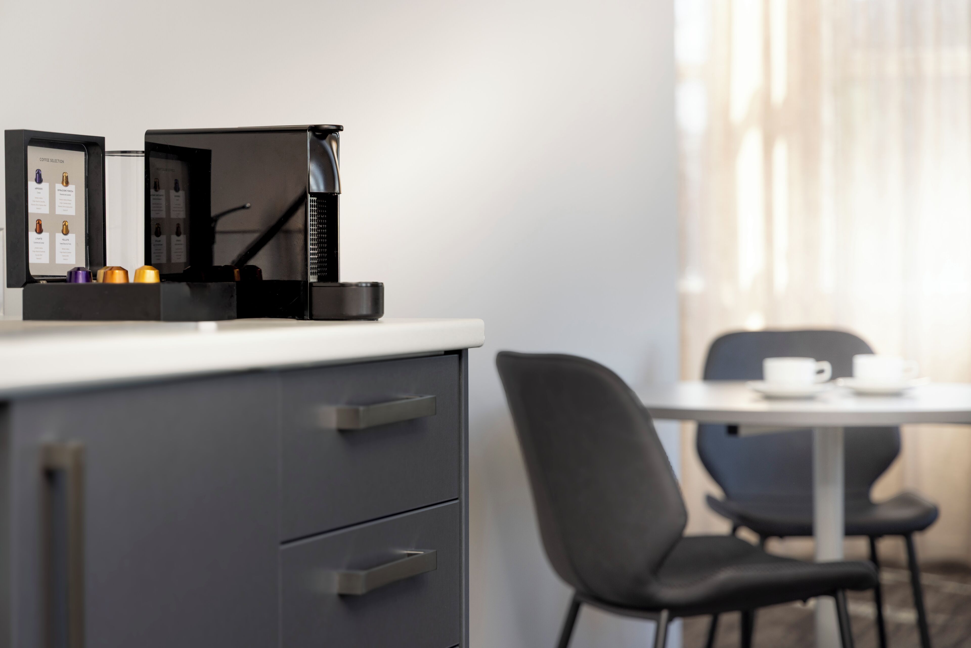 Ein modernes Hotelzimmer mit Kaffeemaschine, einer kleinen Kommode, einem Tisch mit Tasse und einem Stuhl.