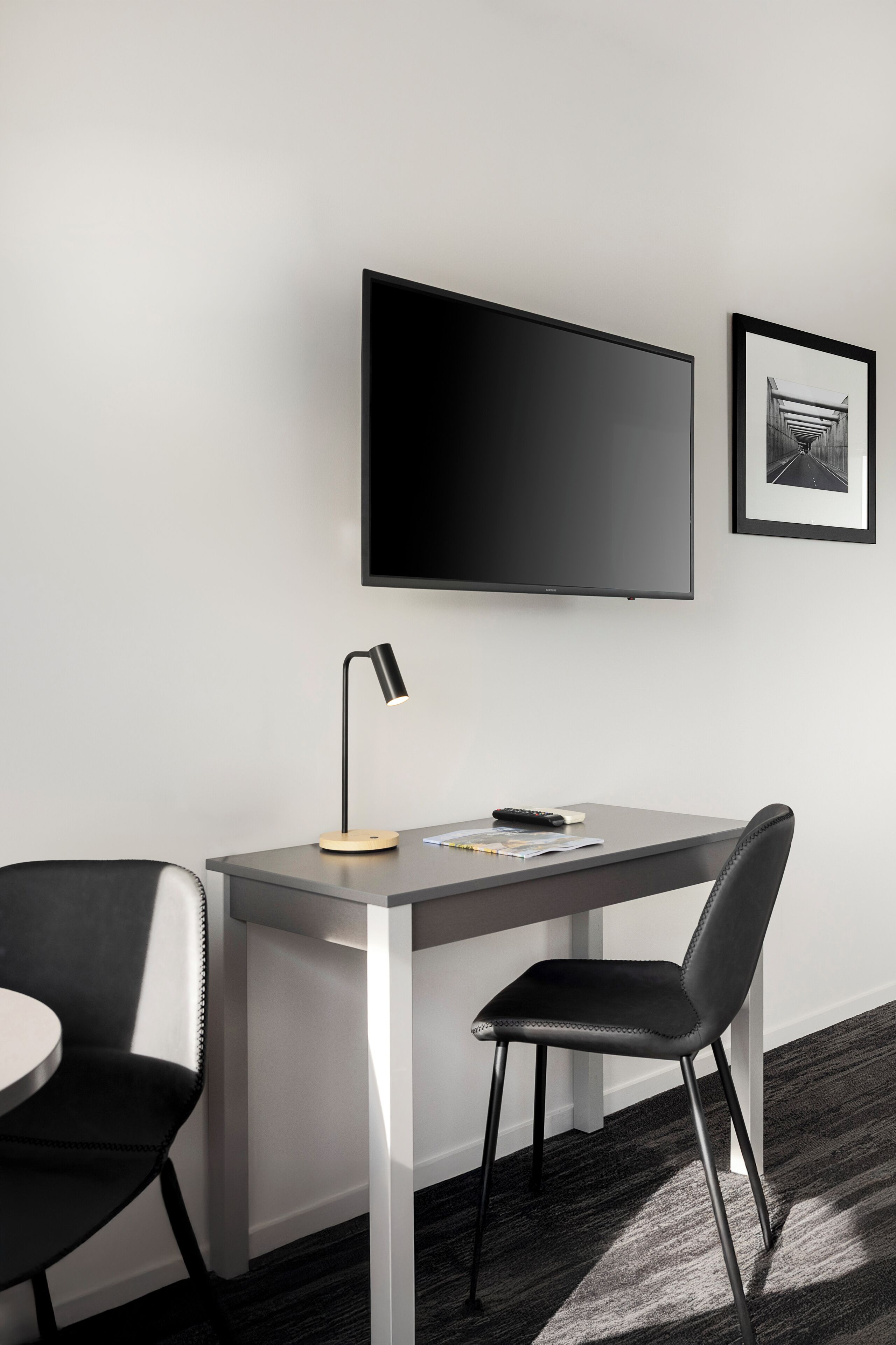 A modern room with a desk, two chairs, a flat-screen TV, a framed picture, and a desk lamp.