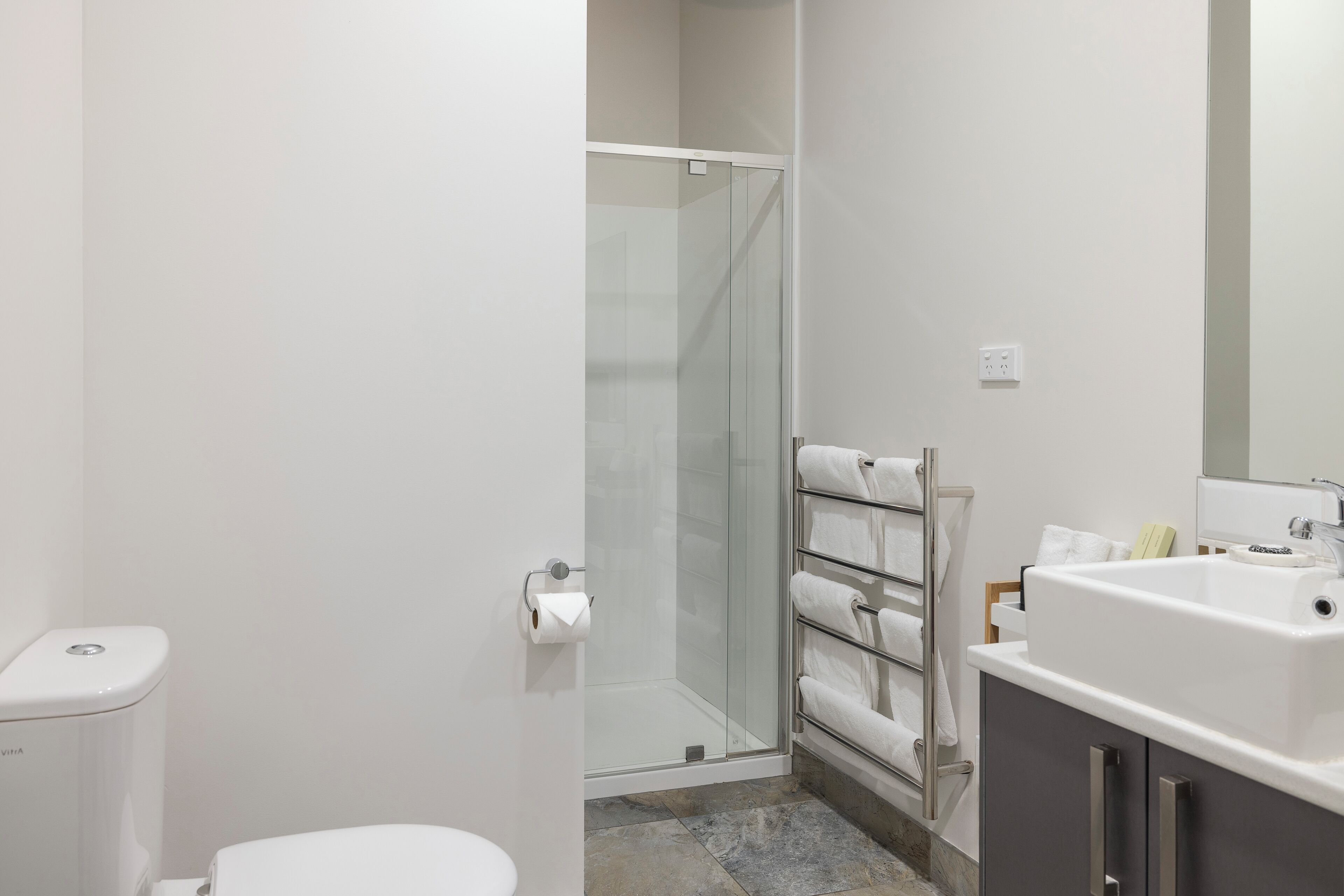 Et moderne badeværelse med glasvæg omkring brusenichen, toilet, håndvask med spejl og bøjle til håndklæder.
