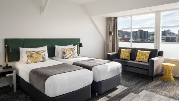 A hotel room with two beds, a sofa, a small table, and a large window offering a city view.