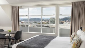 A hotel room with a large window, a bed, a desk with a chair, and a small table with a bowl of fruit.