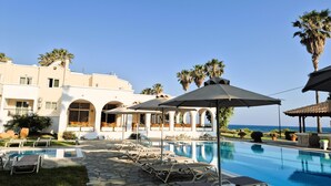 Zona de piscina con sillones de descanso y sombrillas, vista despejada del mar y un edificio con ventanas arqueadas.
