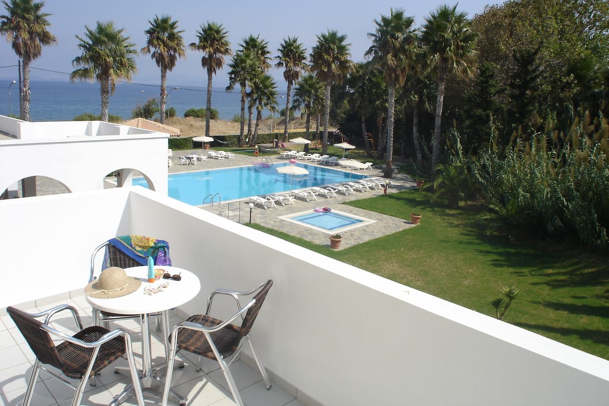 Ein Poolbereich mit Liegestühlen und Sonnenschirmen, umgeben von Palmen und Blick aufs Meer.