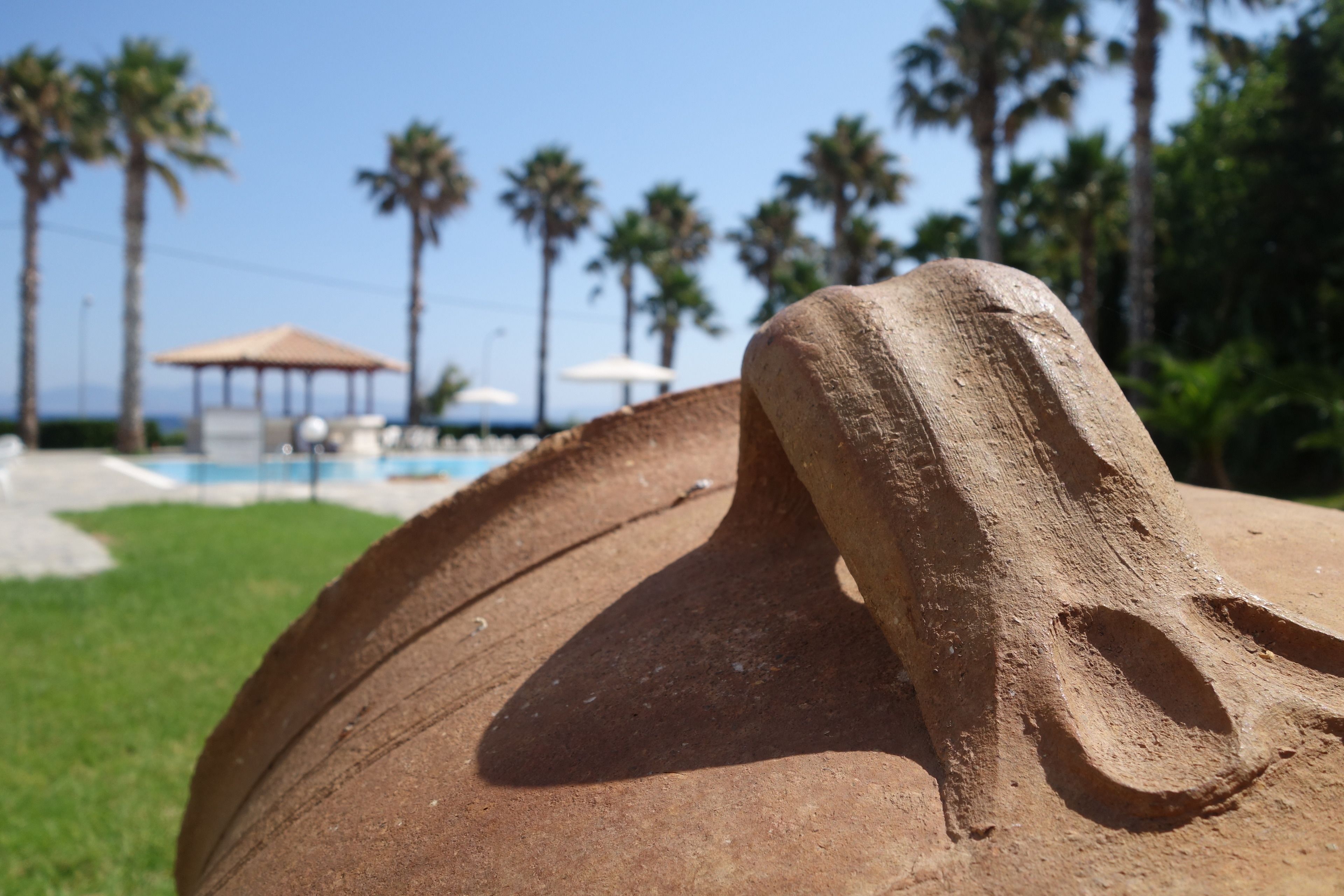 A close-up of a terracotta sculpture with a curved handle, set against a backdrop of palm trees and a clear sky.