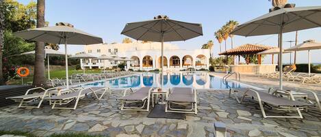 A pool area with lounge chairs and umbrellas, a stone-tiled deck, and a building with arched windows in the background.