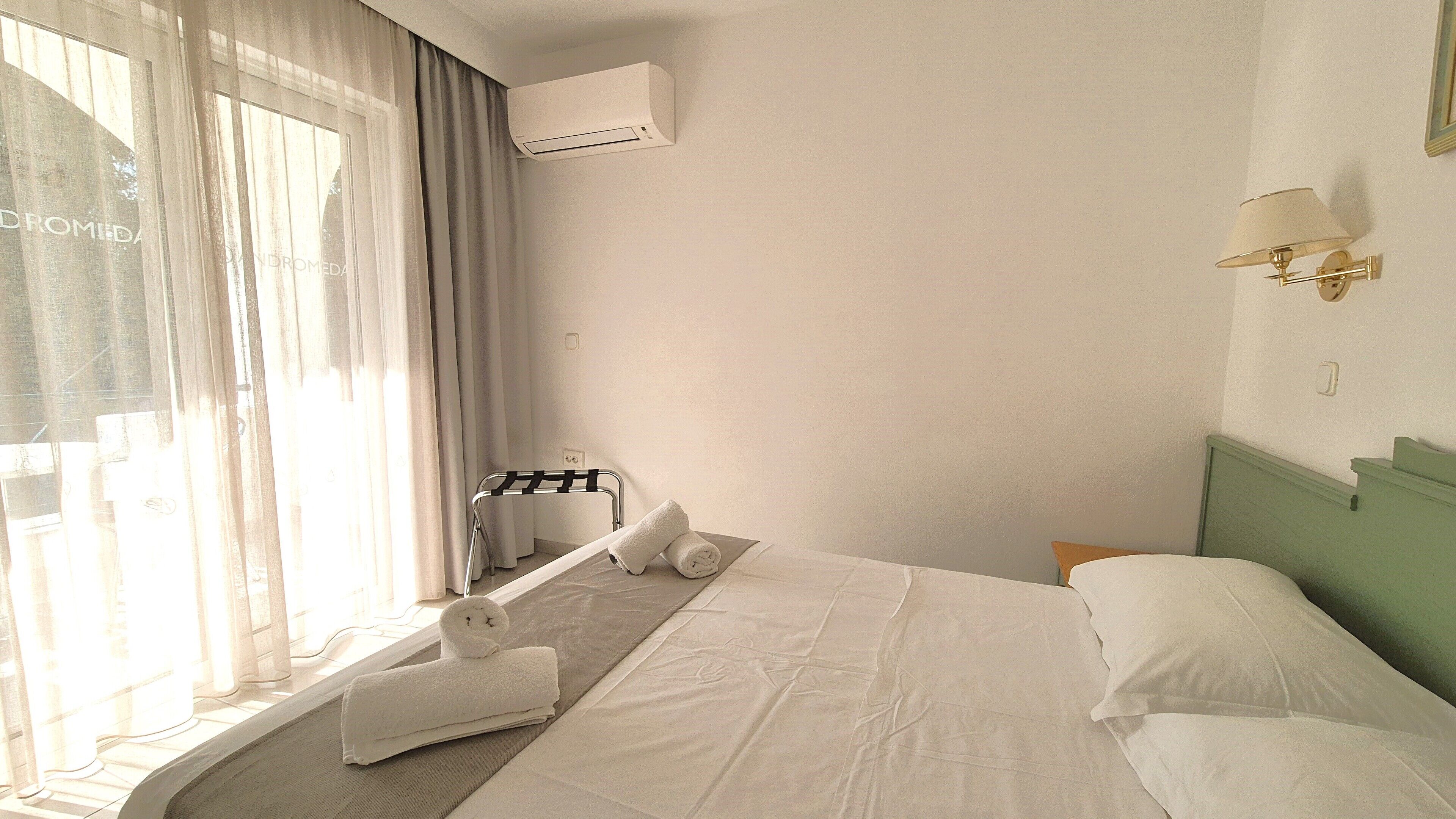 Een hotelkamer met een bed, een raam met gordijnen, een airconditioning, een handdoekrek en een wandlamp.